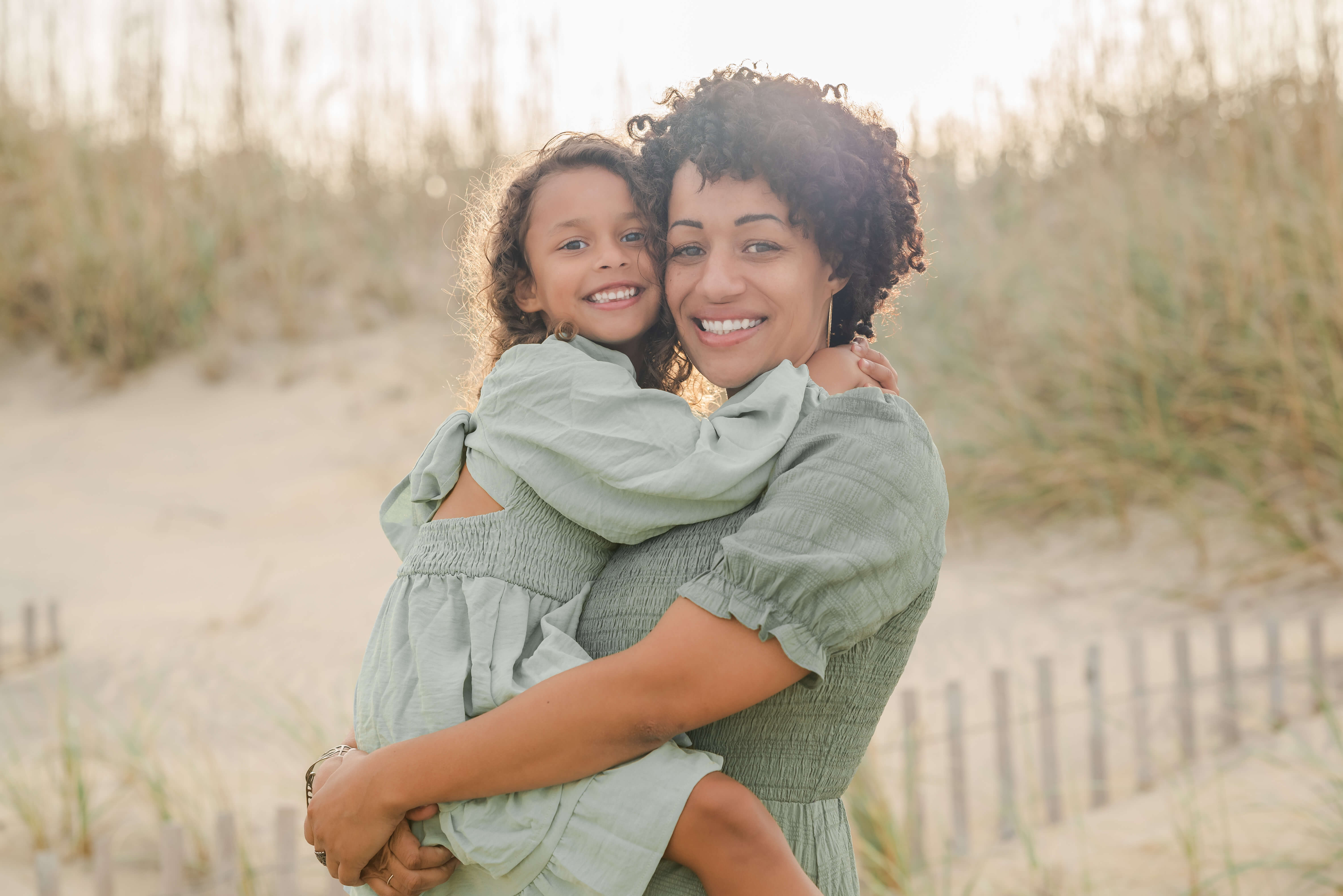 An African American mother in a green dress, holds her young daughter, also in a green dress, in her arms. They are standing in front of the dunes on the beach and the sun is setting behind them.