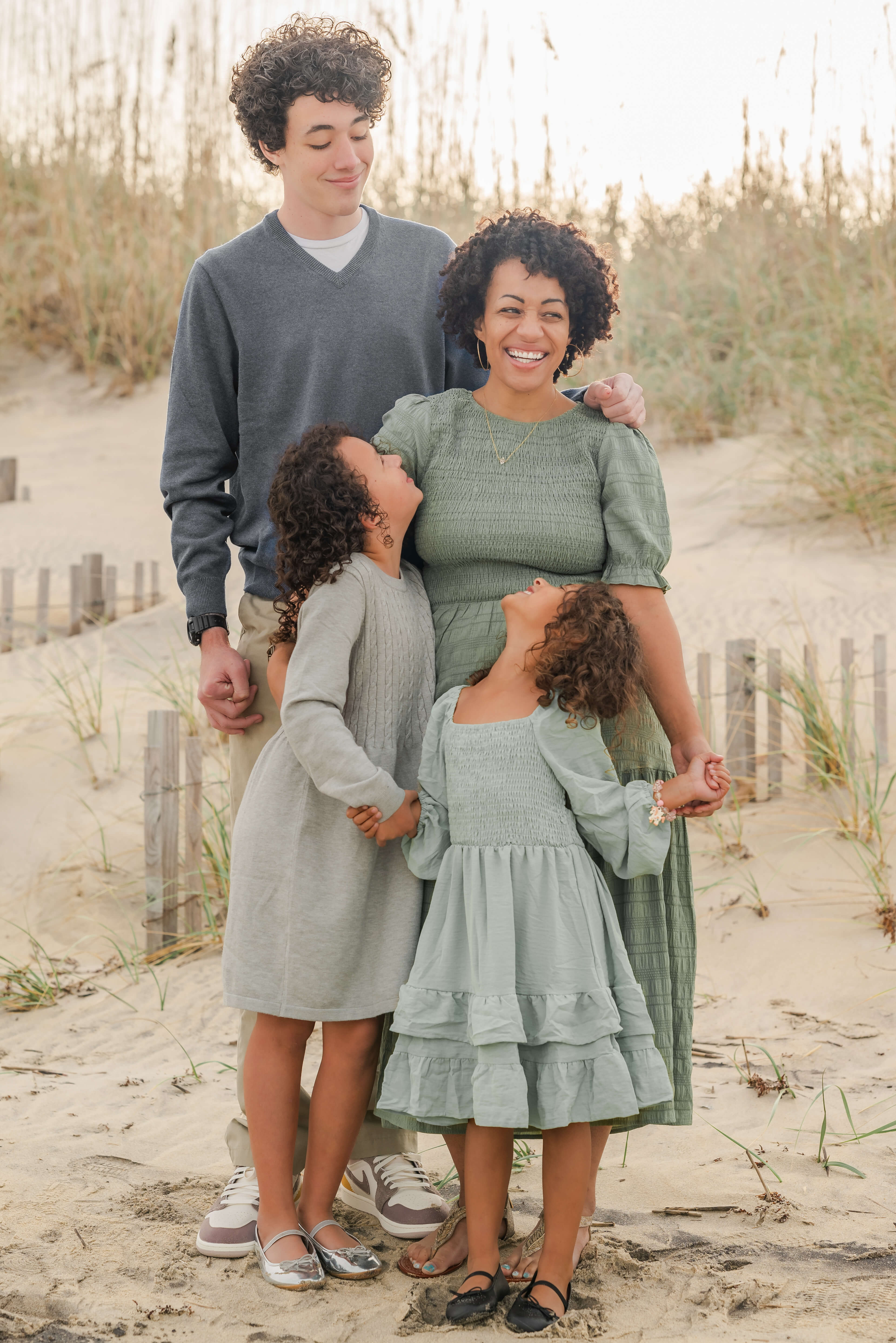 A mother in a green dress laughs while all three of her kids look at her. Her son, who is much taller, stands in the back. Her young daughters are in front of her.