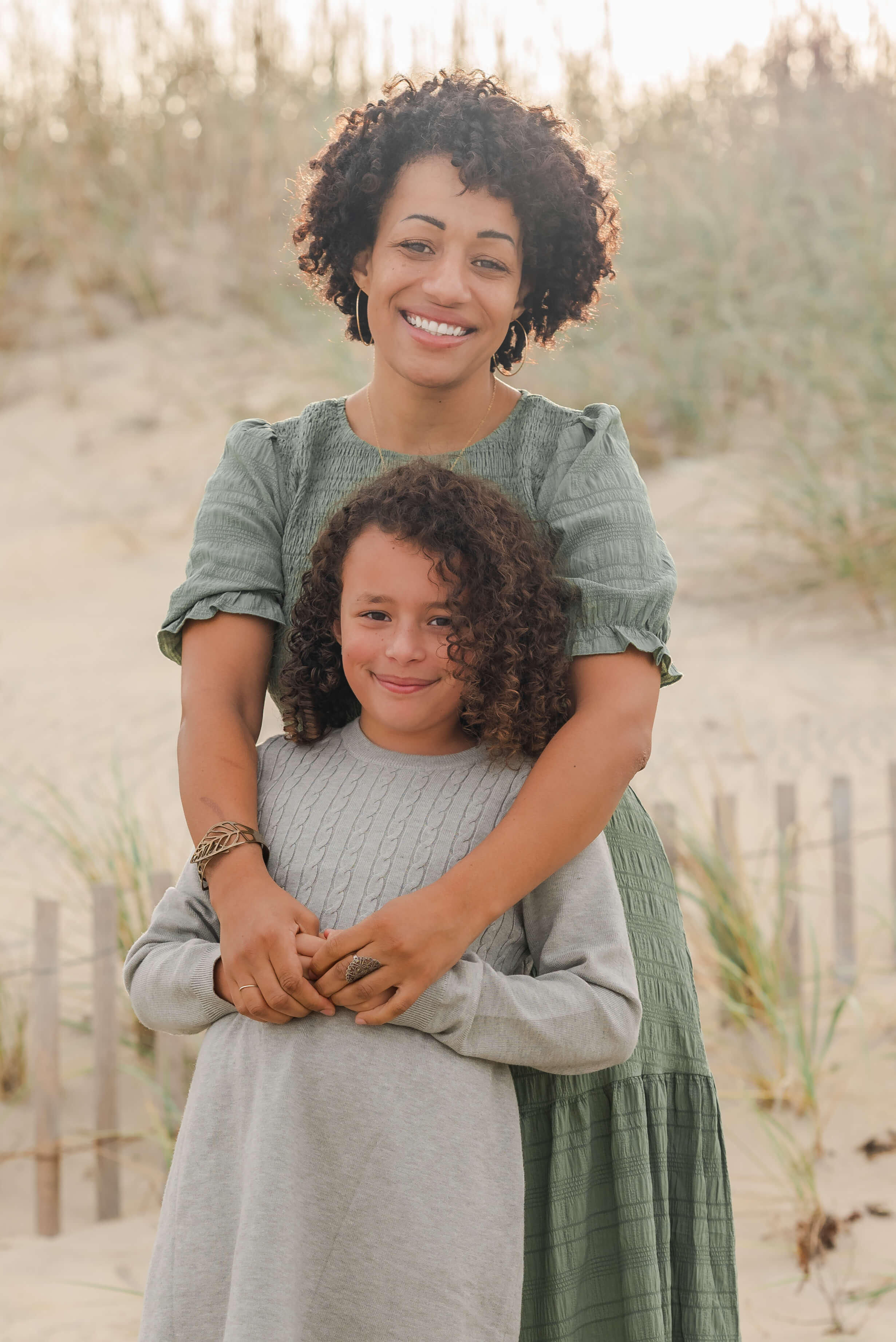 A mother and daughter, both with very curly hair, stand in front of  small fence at the dunes. The mother, in a green dress, stands behind her daughter, in a gray dress, and wraps her arms around her.