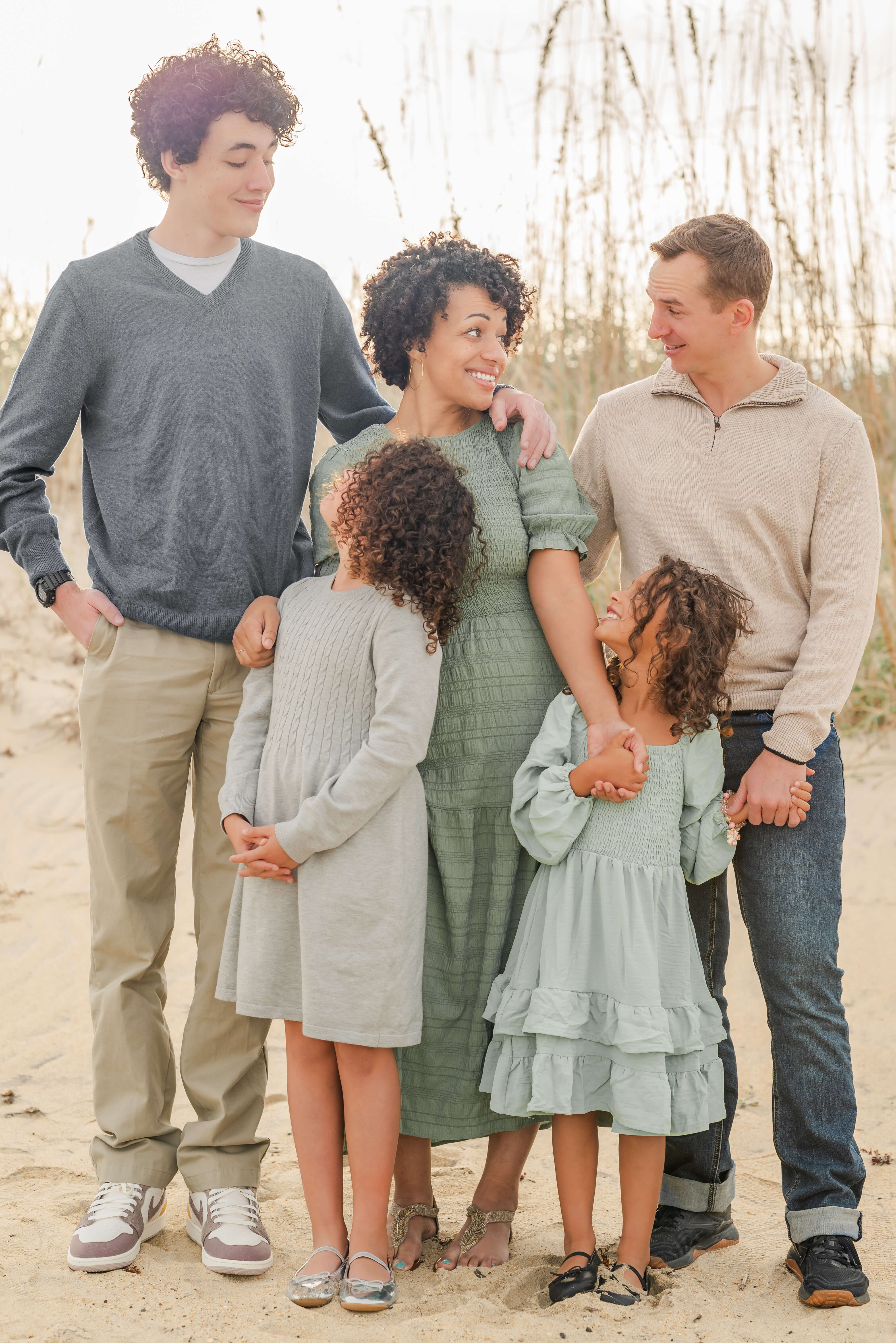 A family of five stand in front of the dunes on the beach. The very tall son, father, and two daughters all look at their mother. The mother looks at her husband.