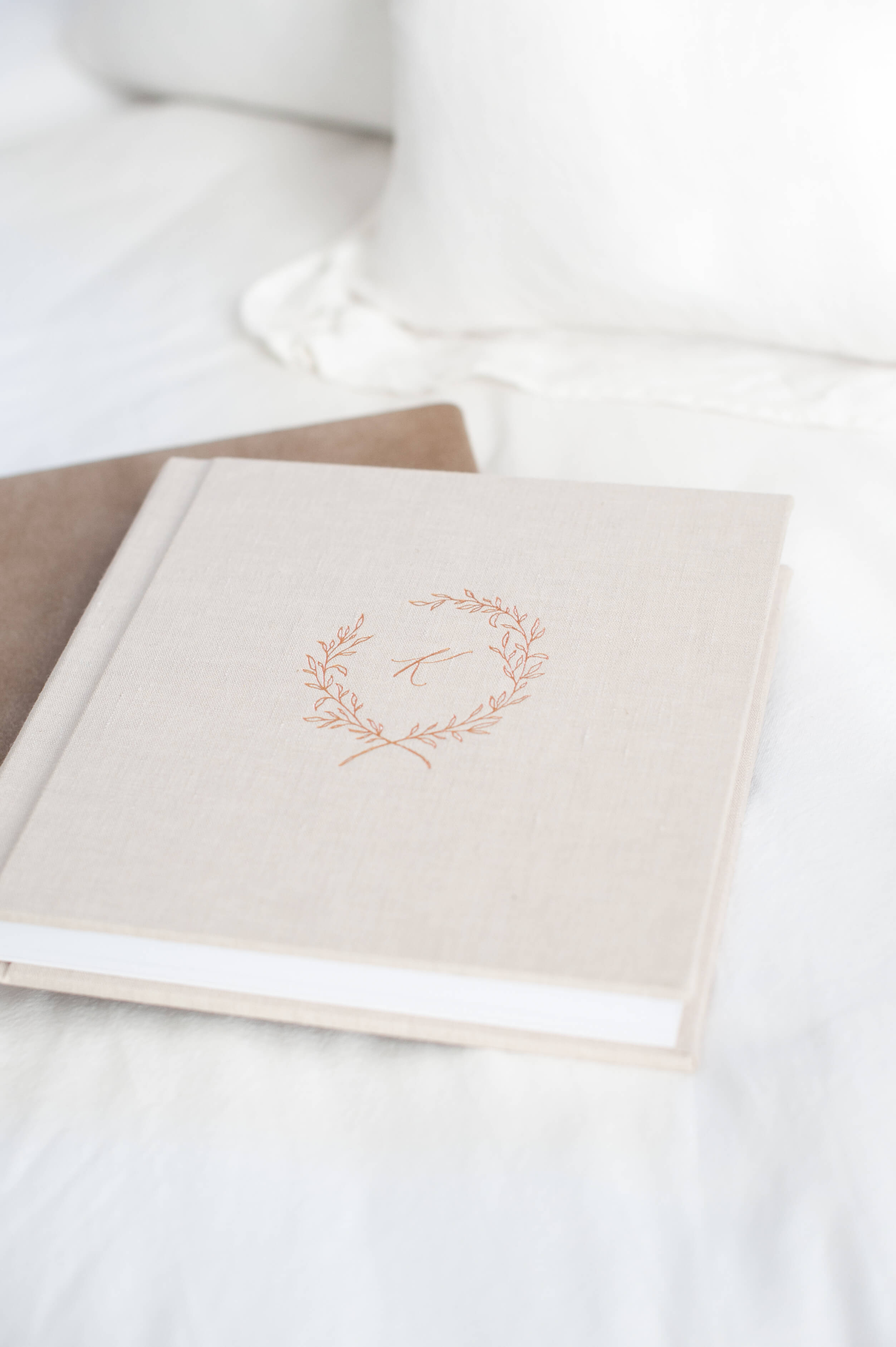 Two heirloom alums sit on a bed with white blankets. The bottom album is brown. The top album is beige linen with copper embossing on the cover.