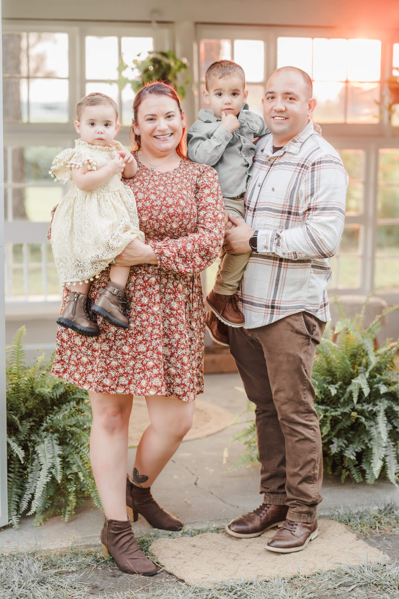 A family of four stands outside of the green house with the sun setting through the windows behind them. They are dressed for a fall family session with Chesapeake photographer, Justine Renee Photography.