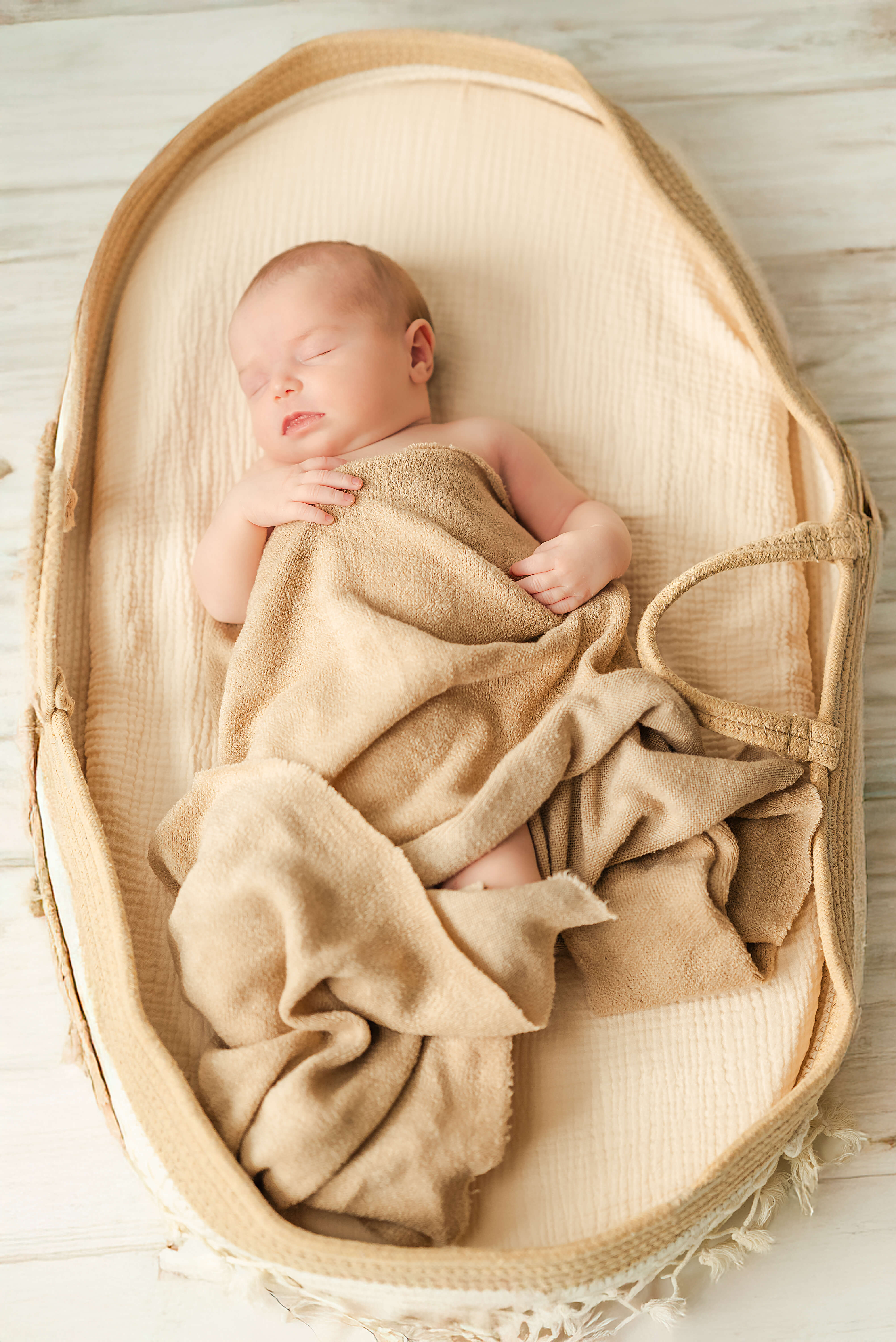 A newborn baby, wrapped in a brown knit blanket, sleeps in a Moses basket on a white wooden floor.