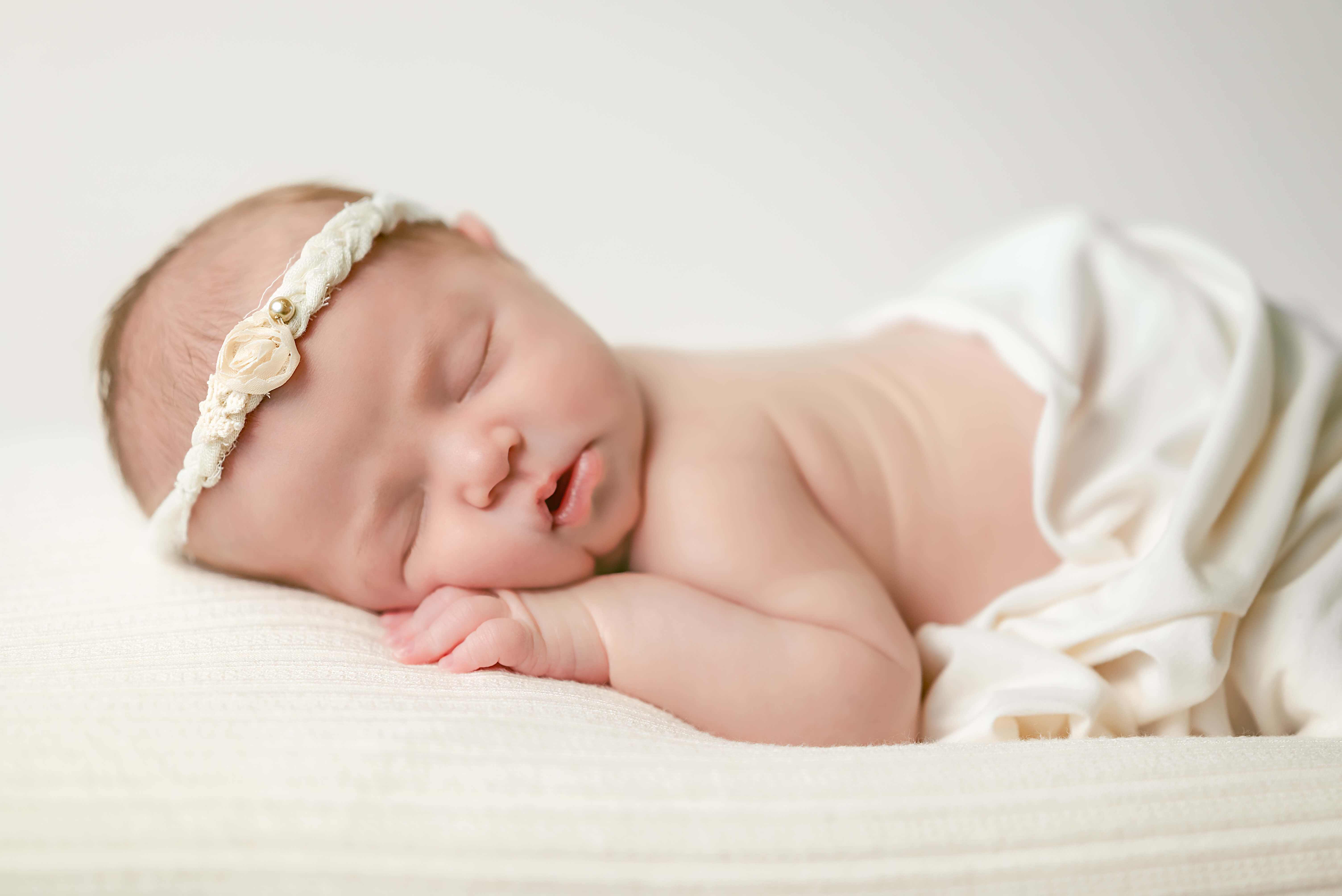 A newborn baby wearing a white floral headband, sleeps on a white blanket. She is draped in another white blanket.