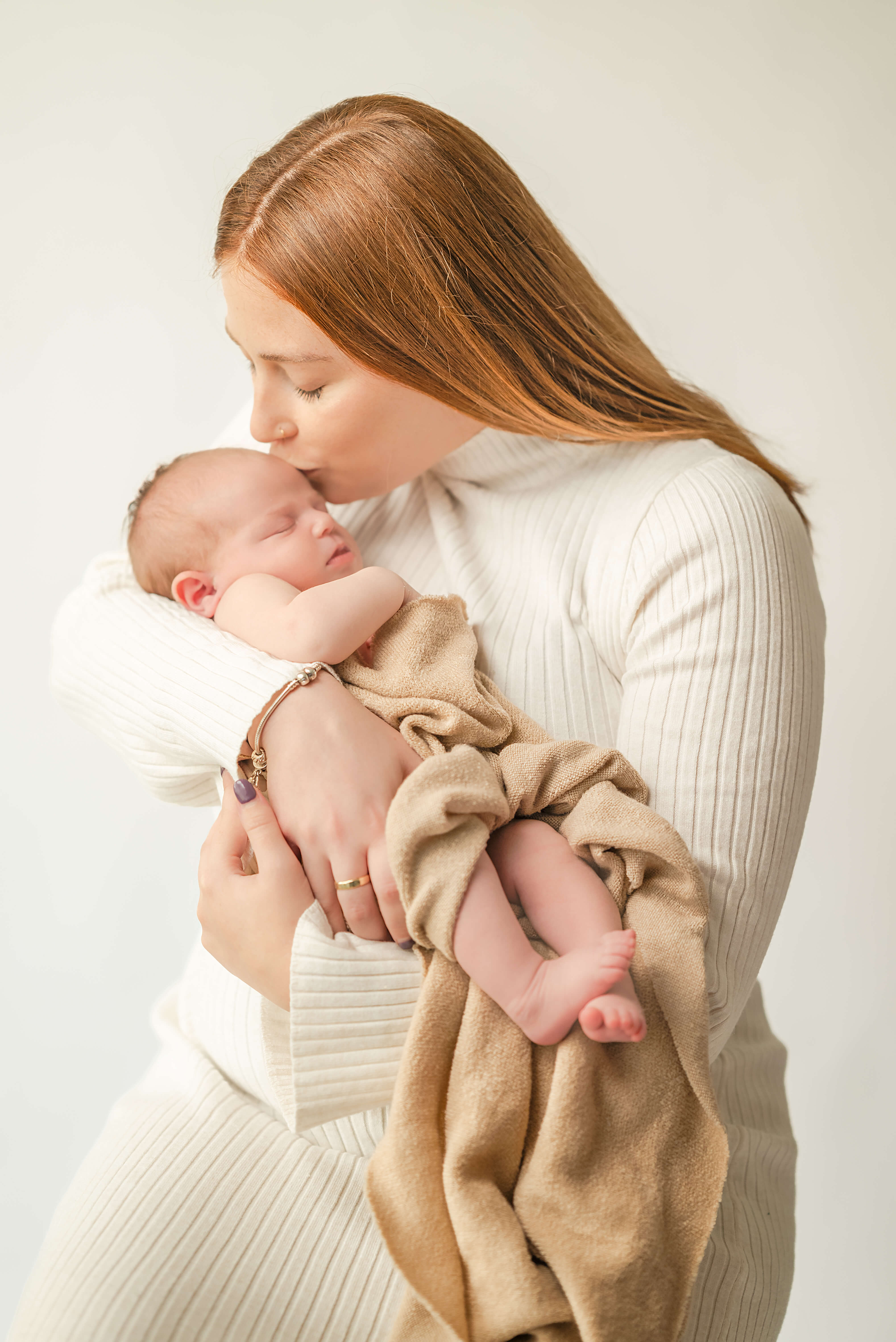  A new mother, wearing an off-white sweater dress with her red hair styled simply, kisses the op of her newborn's head.