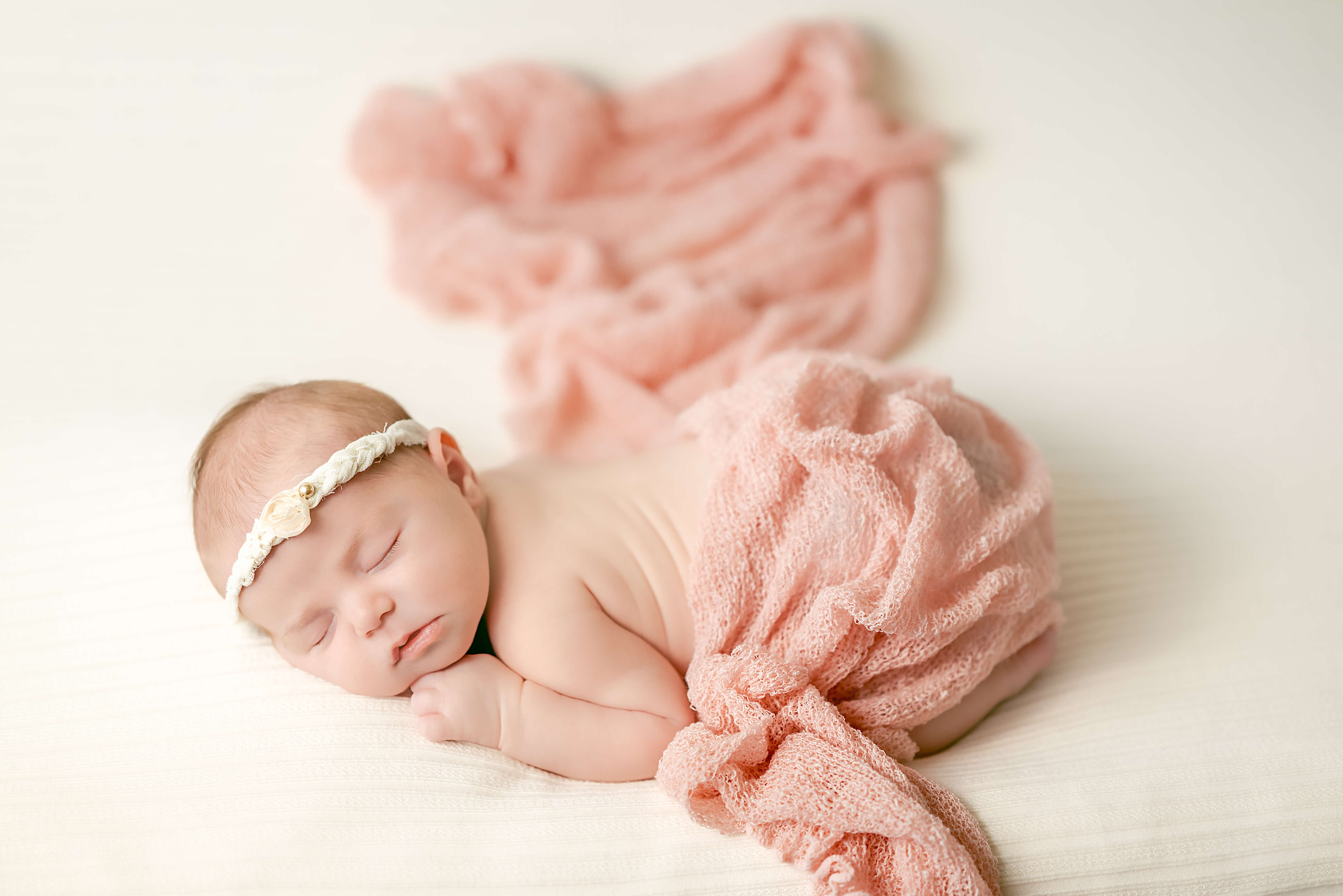 A newborn baby wearing a white floral headband, sleeps on a white blanket. A pink blanket is draped over her and fanned out behind