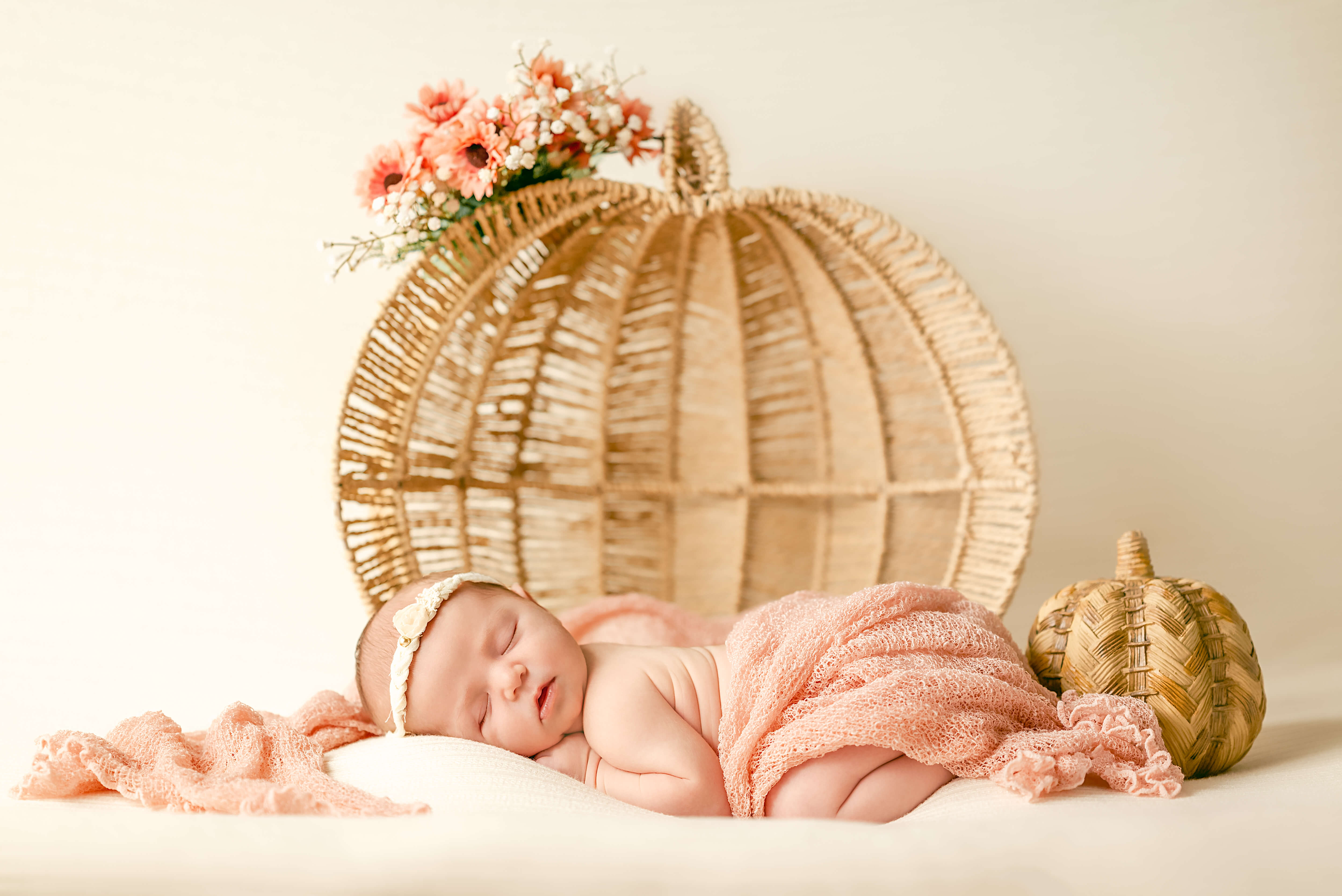 Newborn with a white flower headband sleeps on a white backdrop. She is draped in a pink blanket ad their is a pumpkin shaped basket behind her and a wooden pumpkin off to her side.