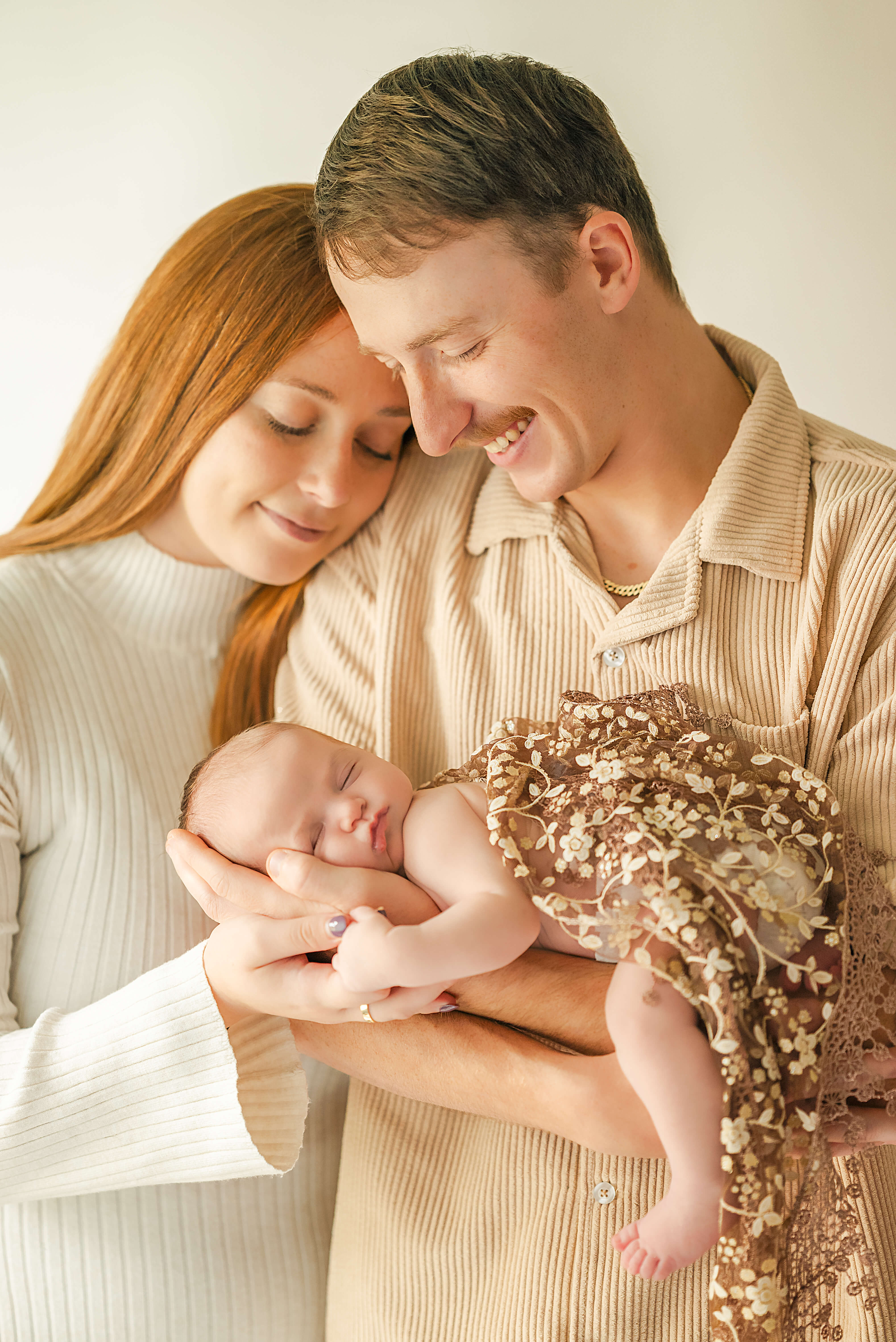 A woman with red hair, wearing a white sweater dress, rests her head on her husband's shoulder. Her husband, wearing a tan shirt, smiles down at their infant daughter, who is wrapped in a floral lace blanket.