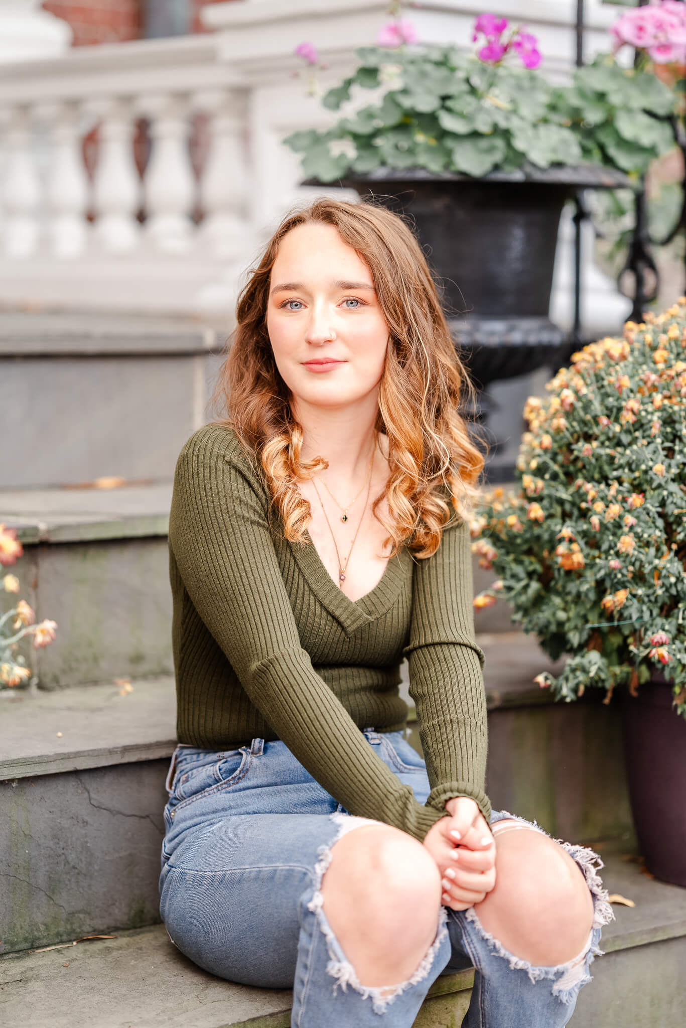 A high school senior wearing a green sweater and ripped jeans sits on some stairs in the Freemason district of downtown Norfolk. She is surrounded by mums and other fall flowers.