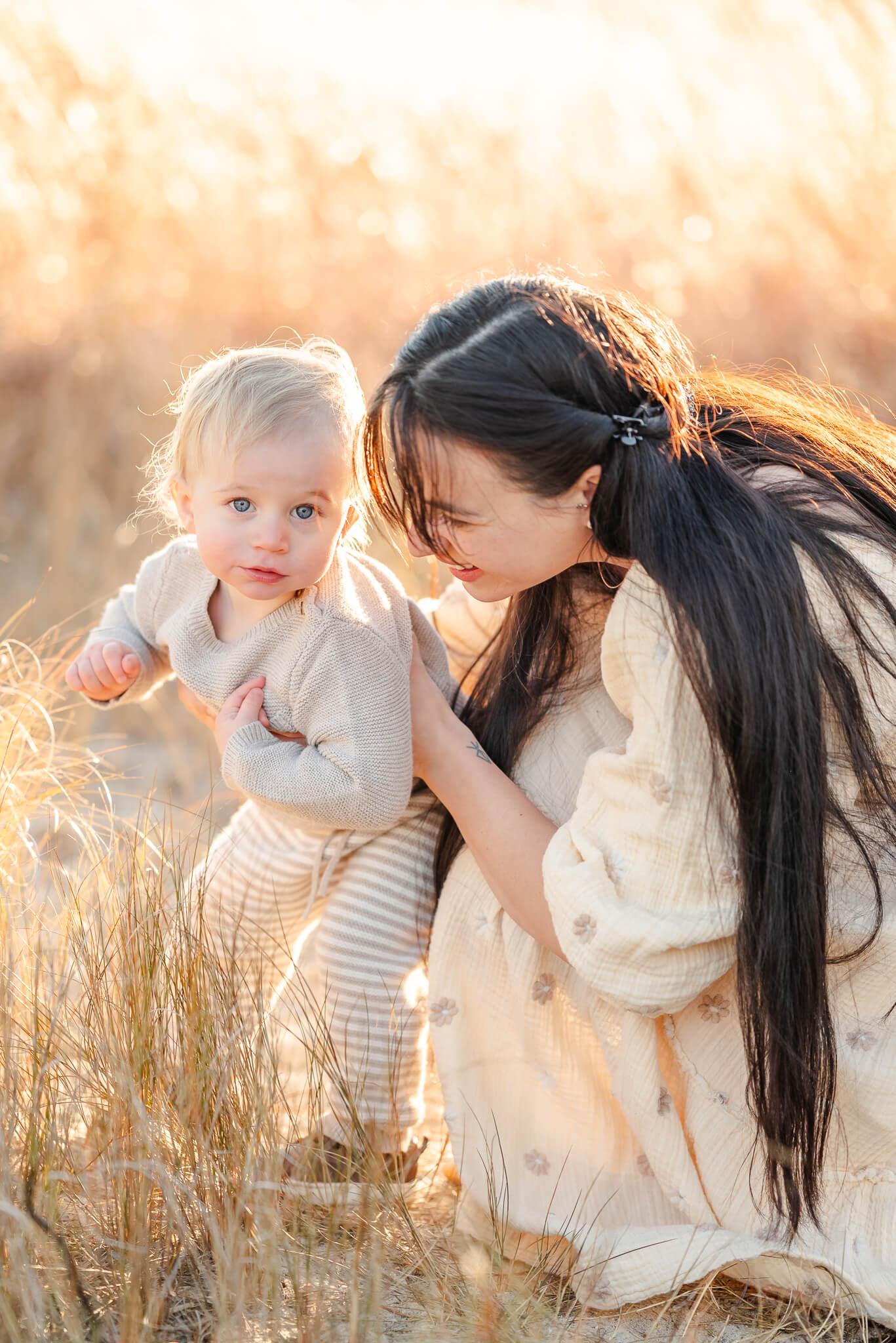 A mother and her toddler son kneel together in some tall brown grass. The mother is wearing a cream dress. The son is in a tan sweater and striped tan pants.