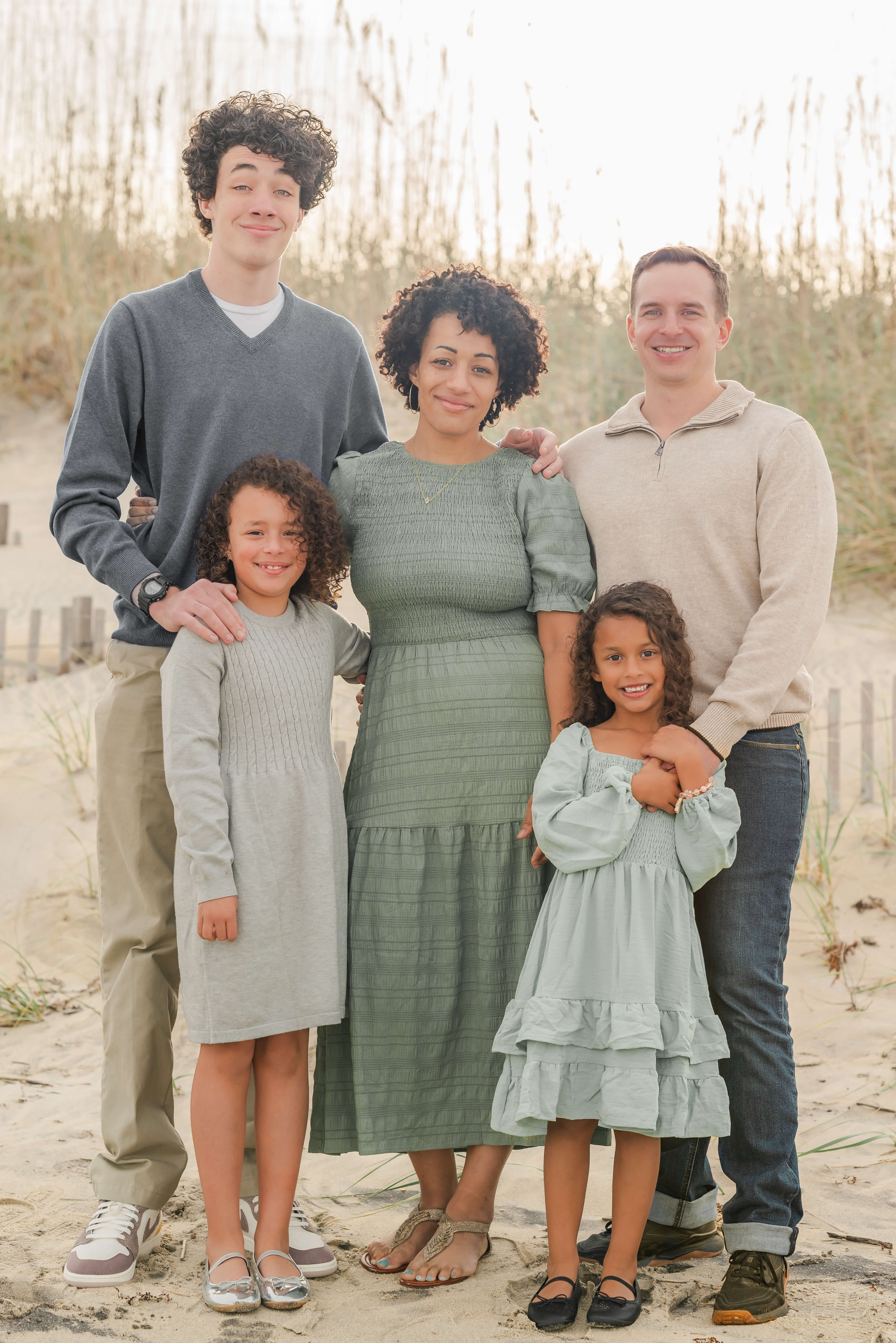 A family of five stands together on Kitty Hawk beach in North Carolina. The mother is in the center in a green dress. Her husband and son stand on either side in tan and blue sweaters. Her daughters, one in a pale green dress and one in a gray dress, stand in front of their father and brother.