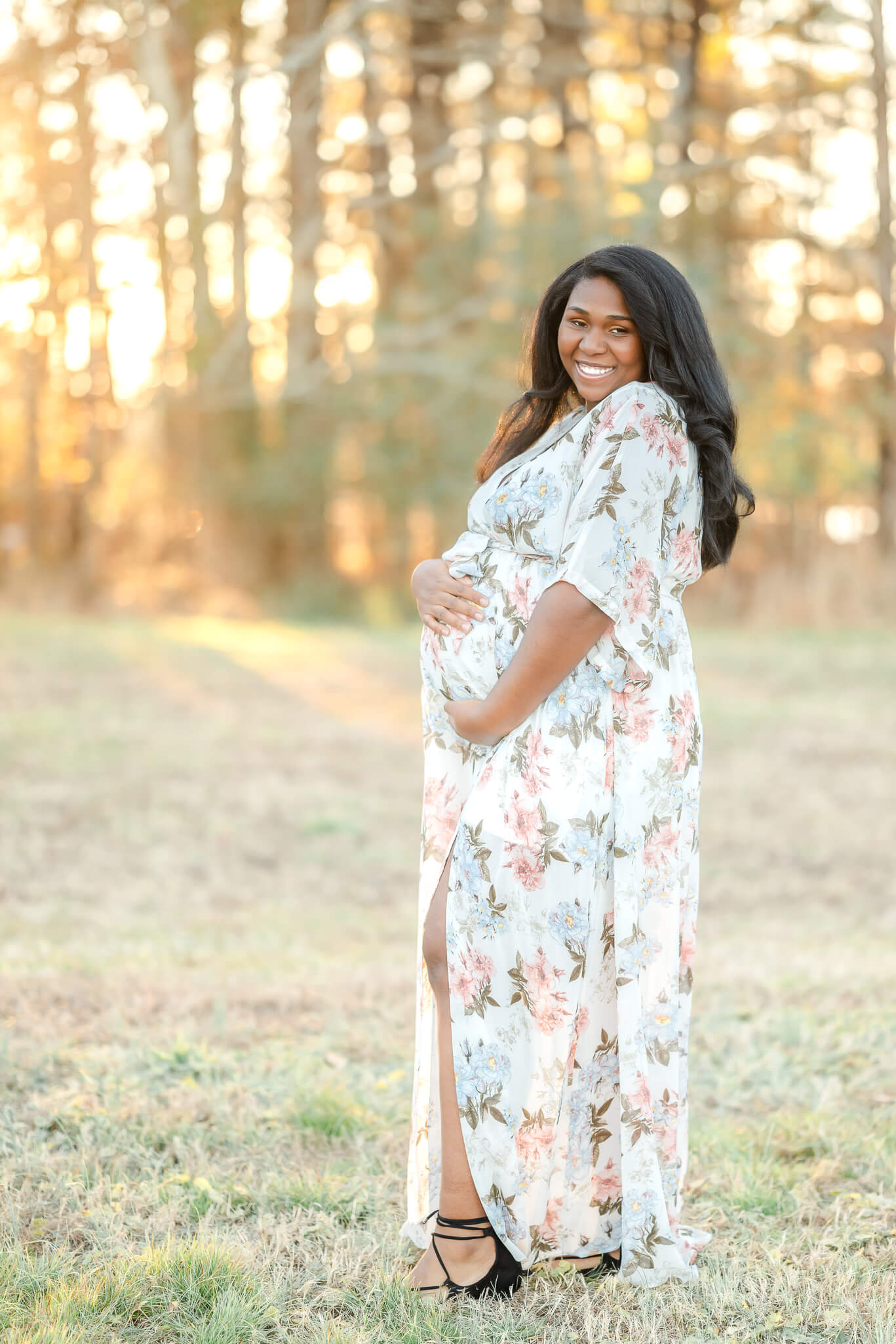 A mother-to-be in a long white, floral dress, cradles her baby bump. She smiles big while the sun shines through the trees behind her.