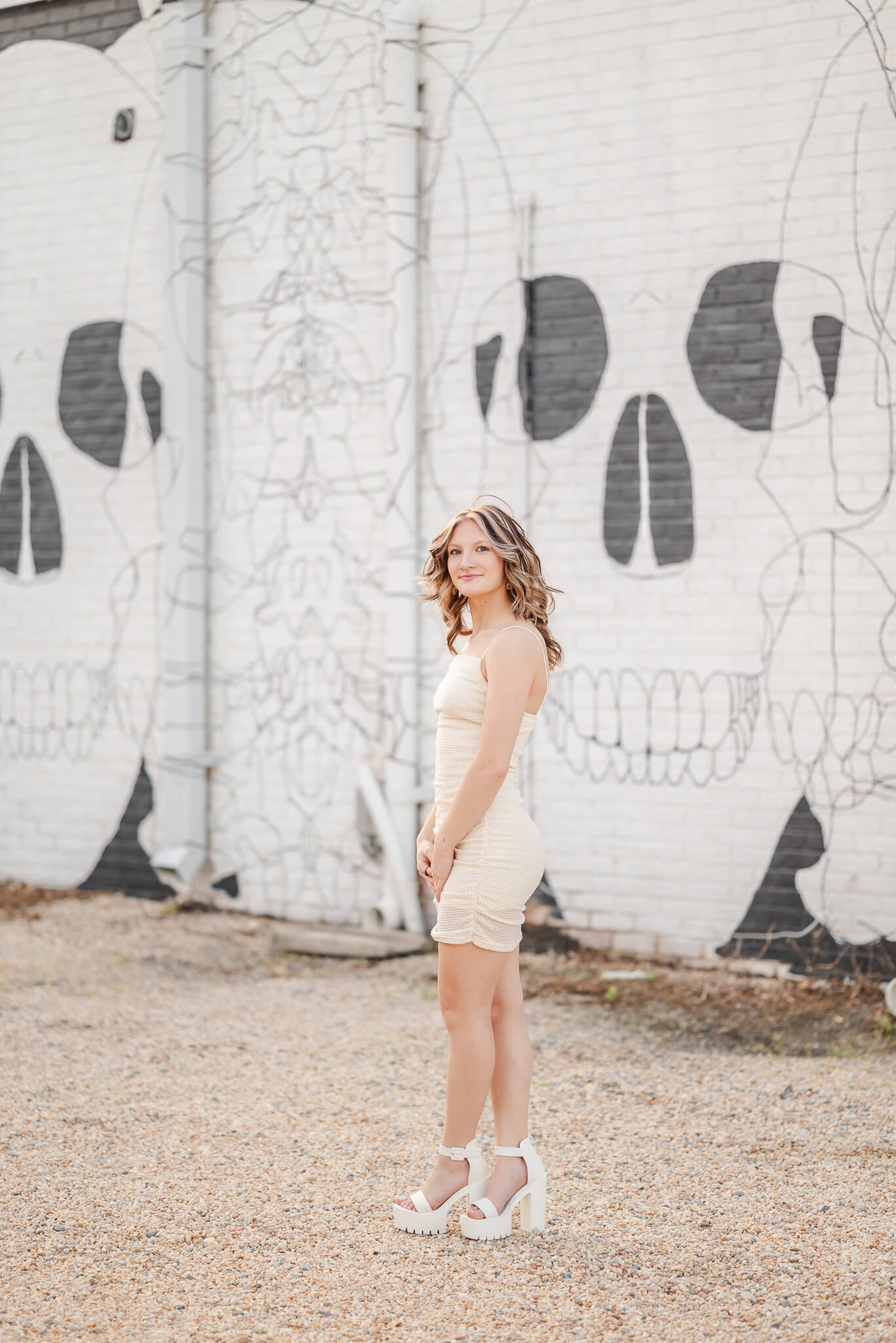 A high school senior, wearing a white bodycon dress and chunky white heels, looks over her shoulder. She is standing in front of a skeleton mural in the Neon district of downtown Norfolk.