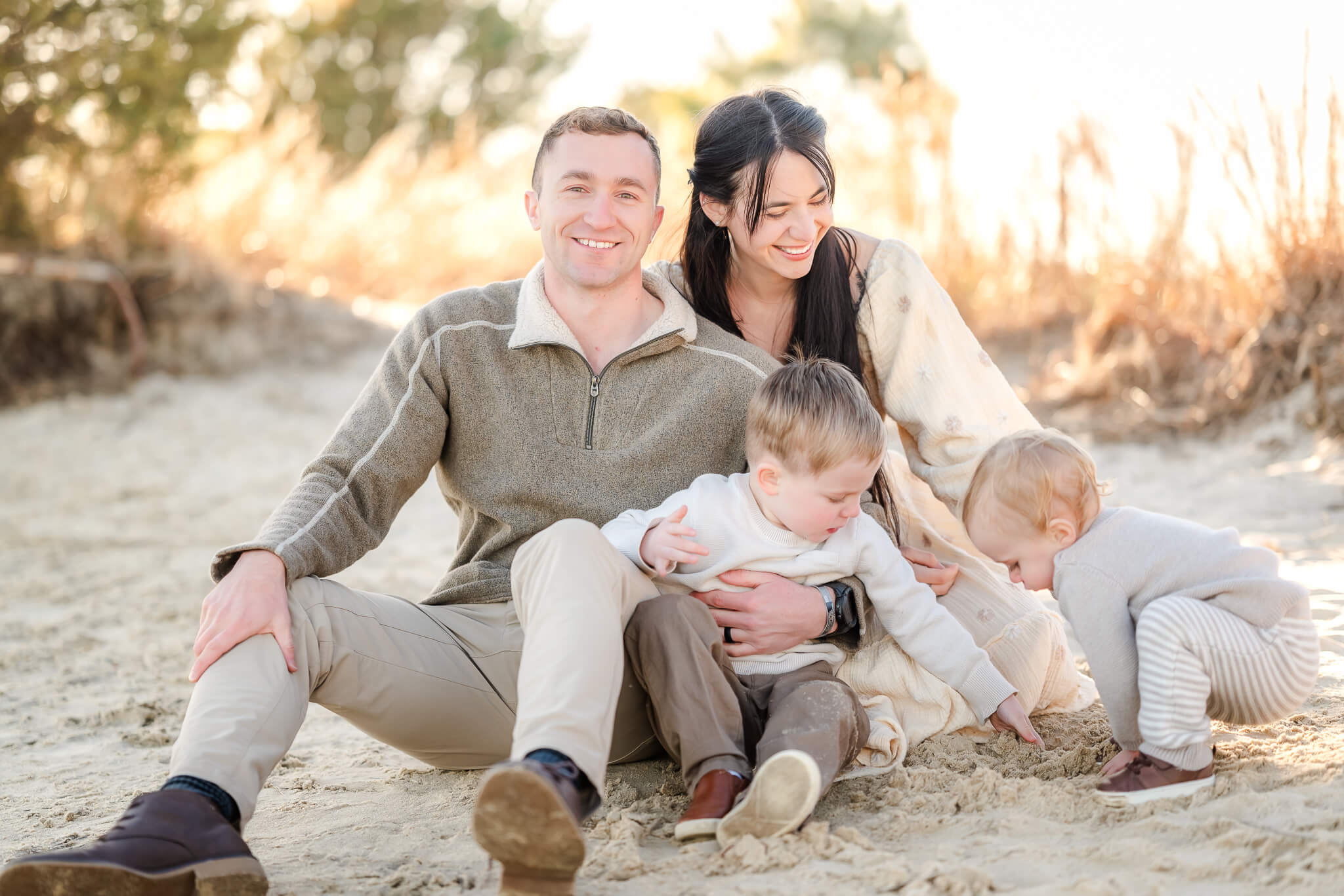 A family of four, all dressed in neutral colors, sits in the and at Pleasure House Point. The toddler boys are both playing in the sand.