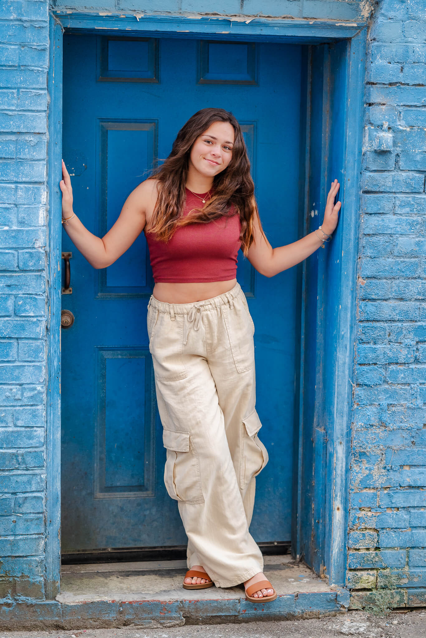 A high school senior wearing loose khaki pants and a red crop drop stand in a doorway. The doorway and surrounding bricks are painted bright blue, fitting for Palin's Alley in downtown Elizabeth City.