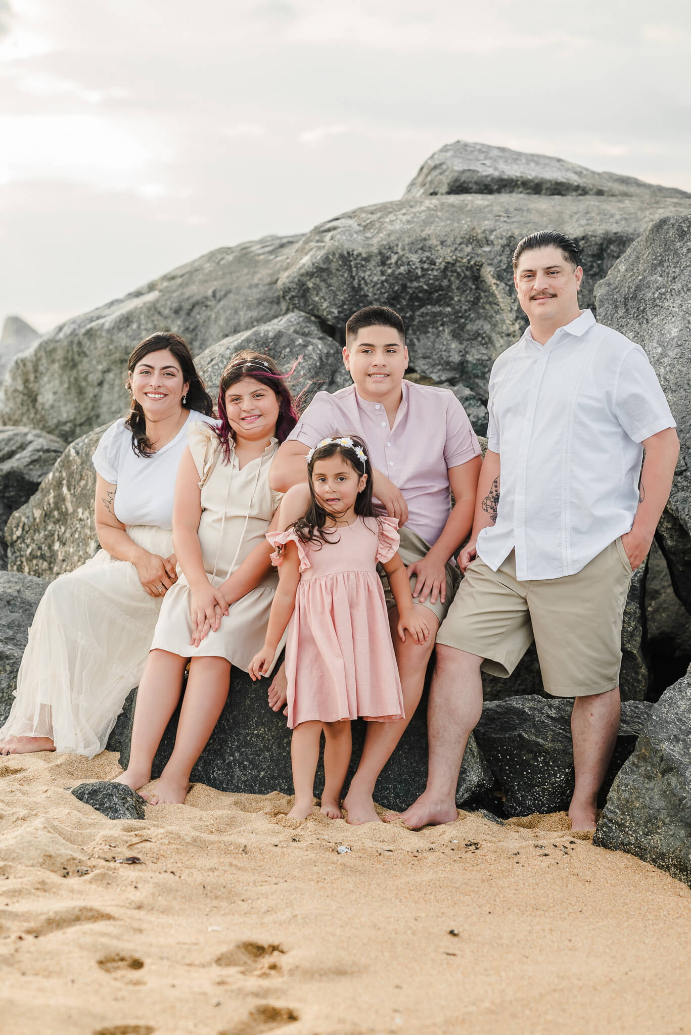 A family of 5 poses on the rocks at Fort Story beach in Virginia Beach. The are wearing a mix of pink, khaki, and white. The parents sit on the outside while the children are clustered in the middle.