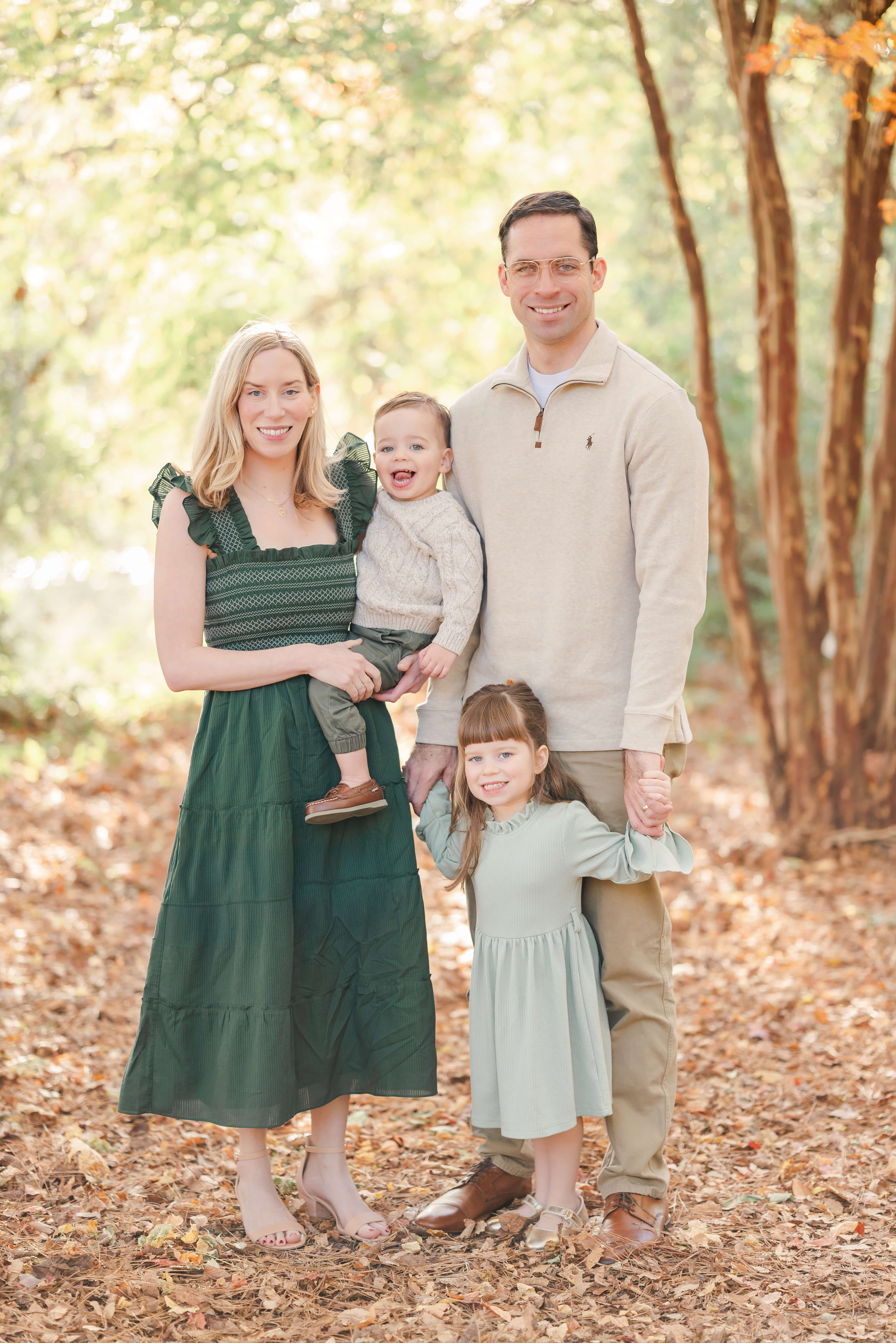 A family of four poses at Oak Grove Lake Park in Chesapeake, VA. The are dressed in greens and tans, which compliments the sun shining through the green leaves behind them. The mother holds the toddler son in her arms. The father holds the hands of the toddler girl, who stands between his feet.