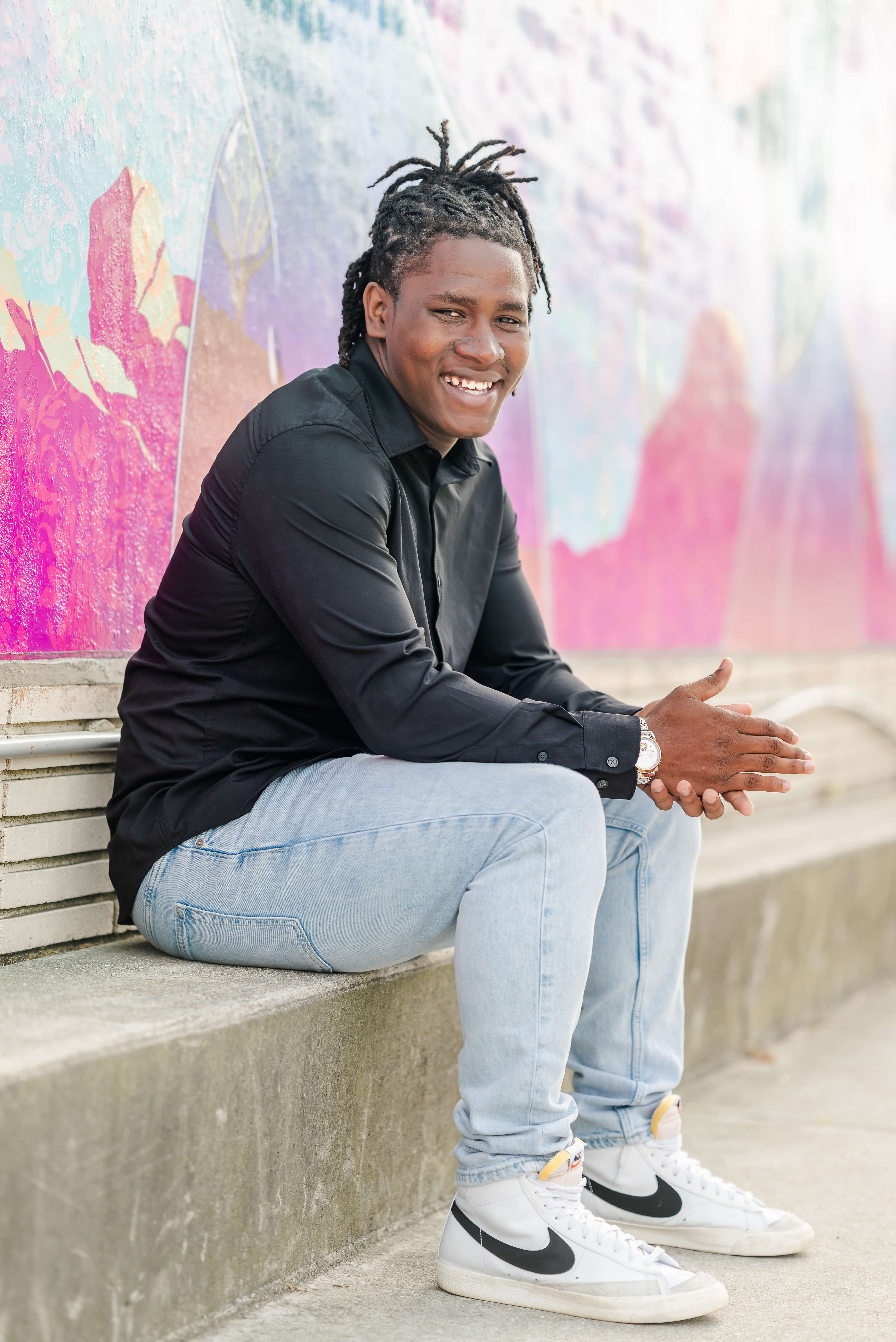 A high school senior in a black shirt and jeans, with his braided hair pulled back, sits on the curb in front of a mural. He is in the Neon District of downtown Norfolk.