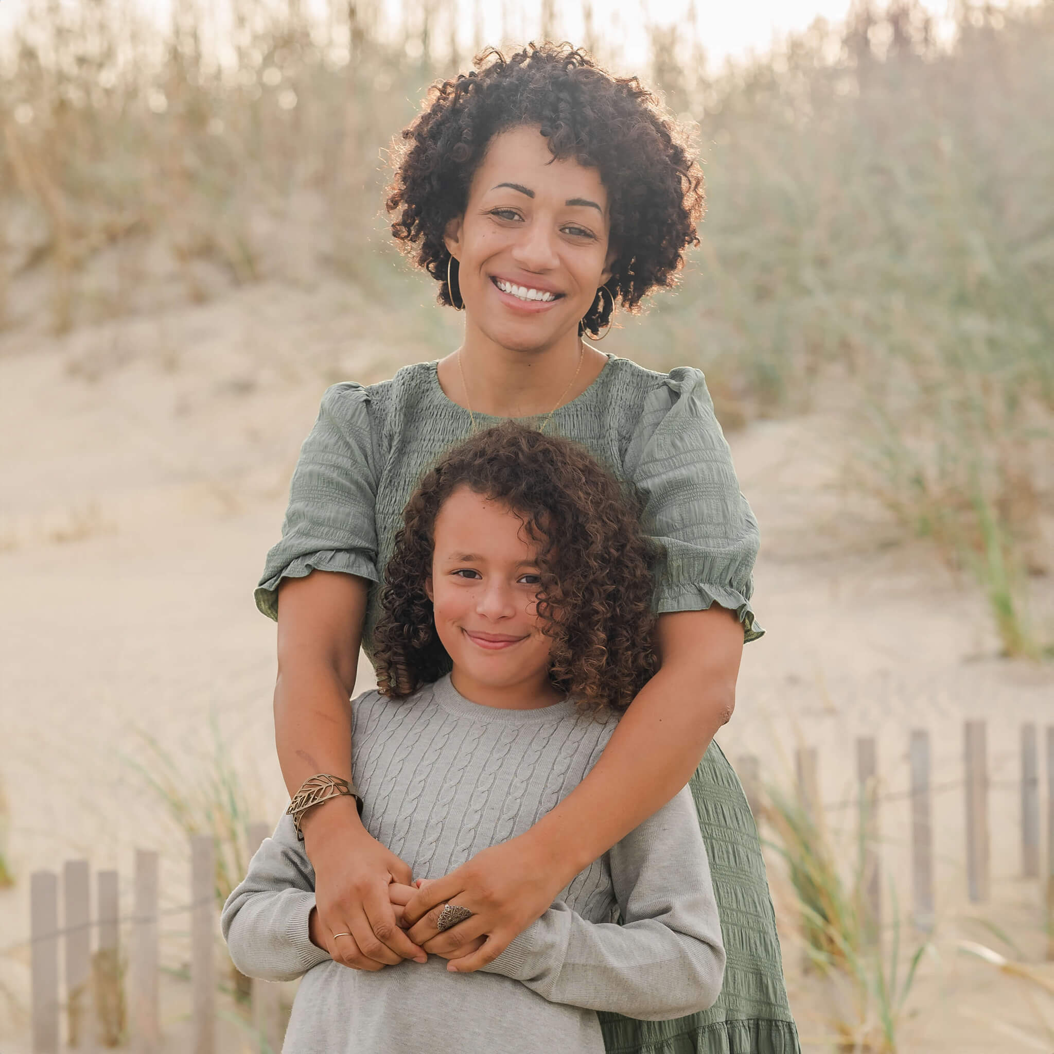 A mother, wearing a green dress, stands behind her daughter who is in a gray dress. She has her arms around her. They are standing near the dunes on the beach with the sun setting behind them.