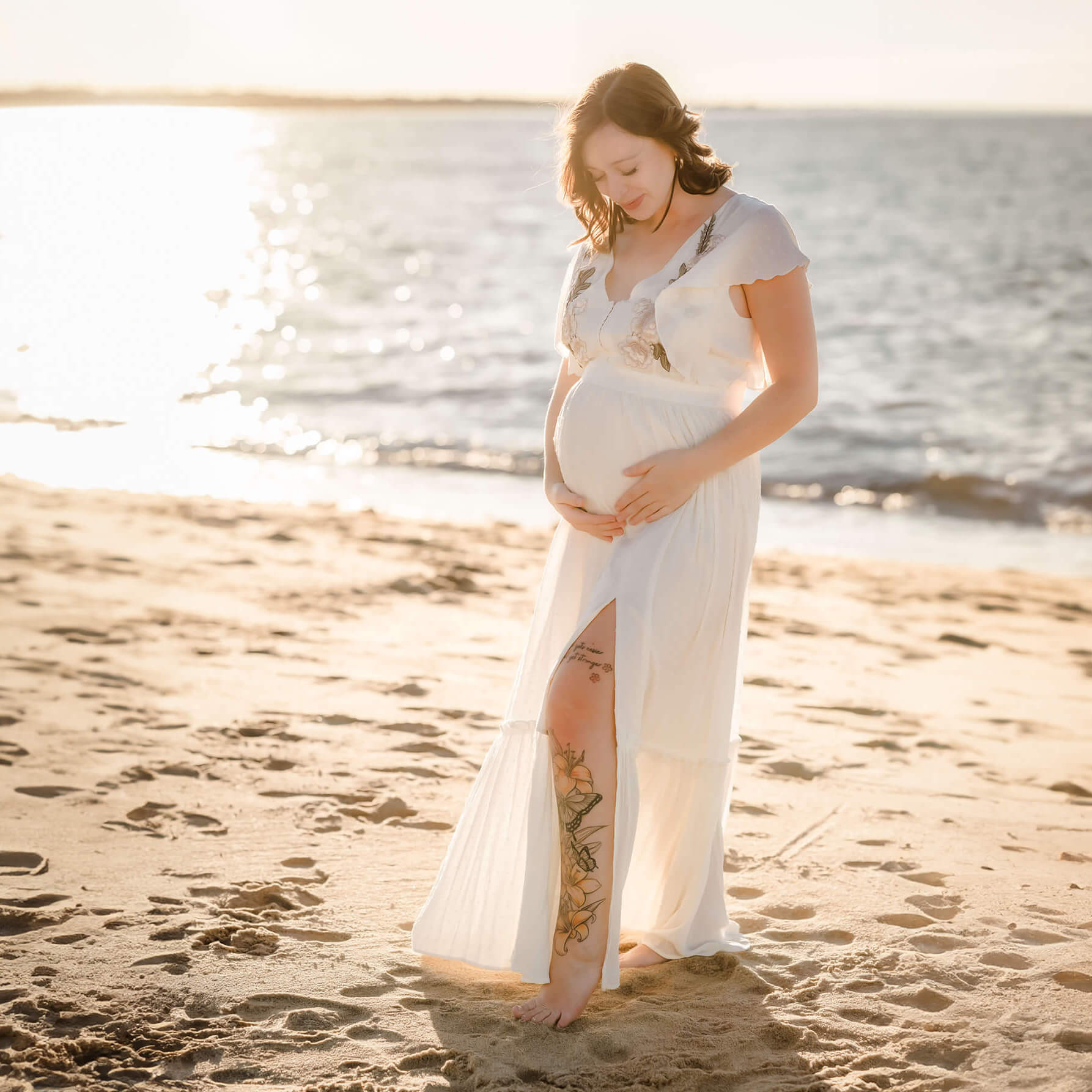 An expectant mother, wearing an off-white dress with a slit, pops her leg and looks lovingly at her belly. The sun is shining off the water and reflecting on the the sand at the beach.