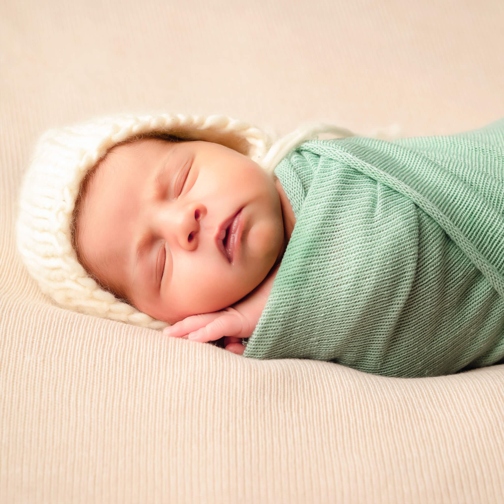 A newborn, with a teal swaddle and white knitted hat, sleeps peacefully on a white blanket.