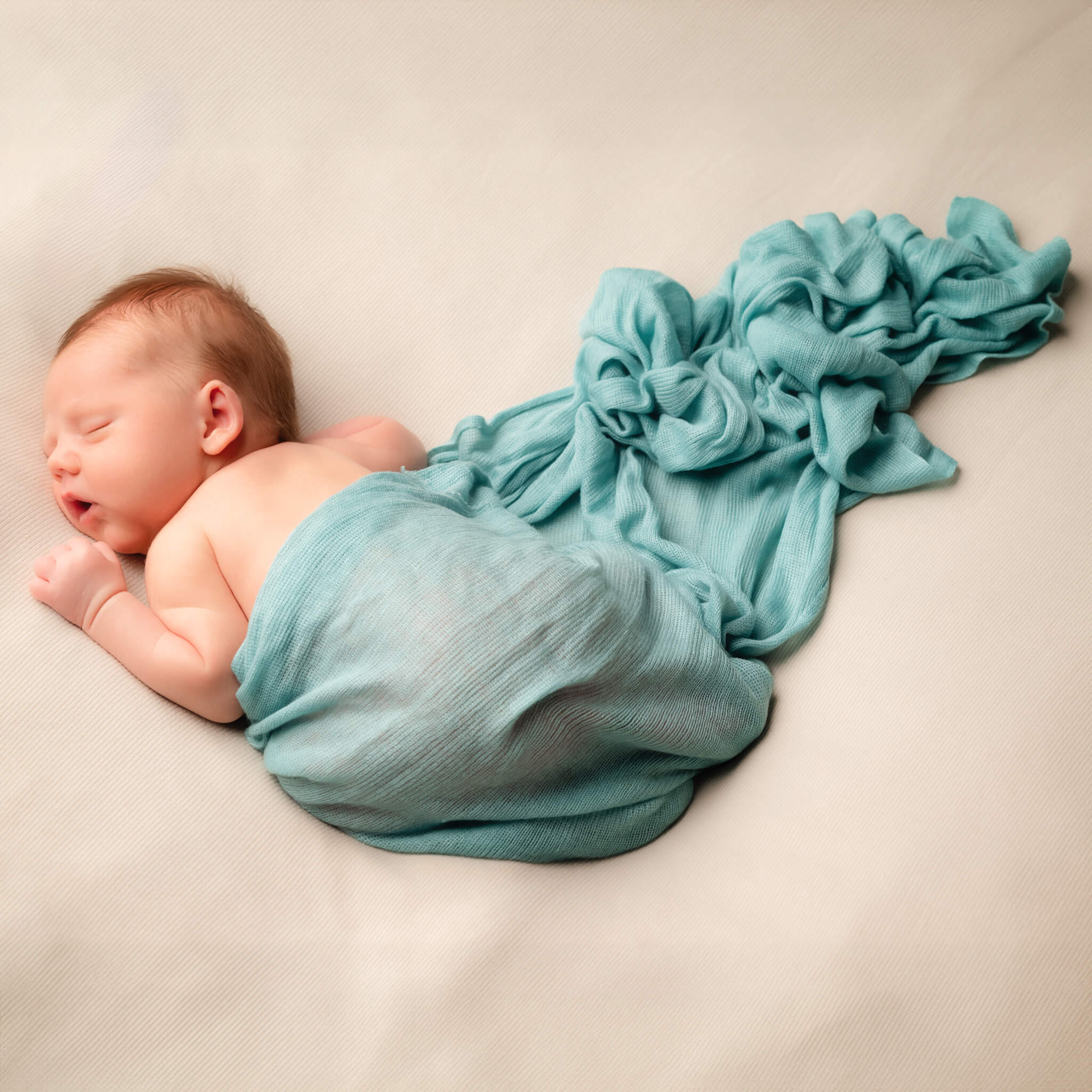 A newborn sleeps on his stomach on a white blanket. A blue blanket is draped over him and fanned out.