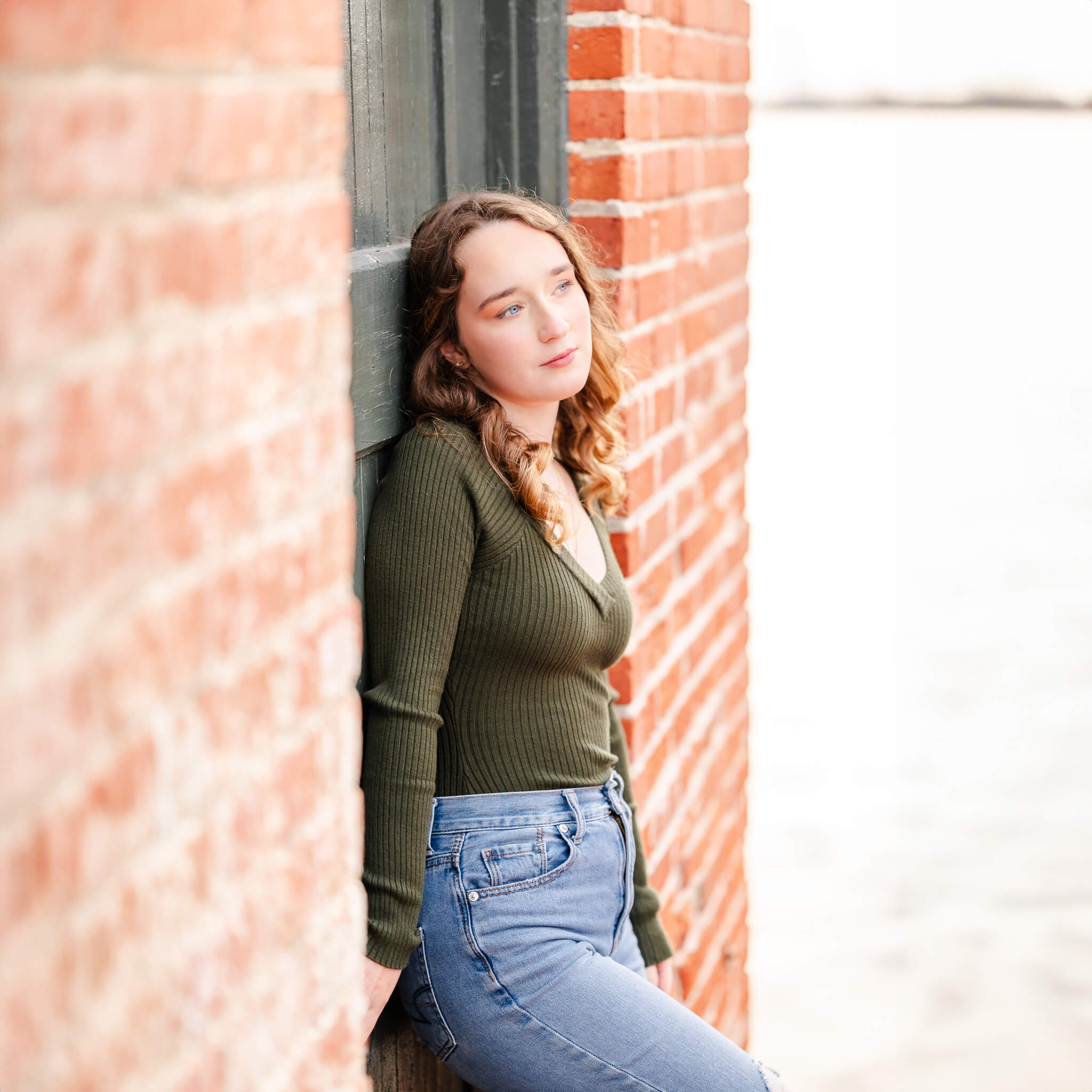 A high school senior, wearing a green sweater and jeans leans against a black door in a brick building. There is water to her left, with a bit of land in the distance.