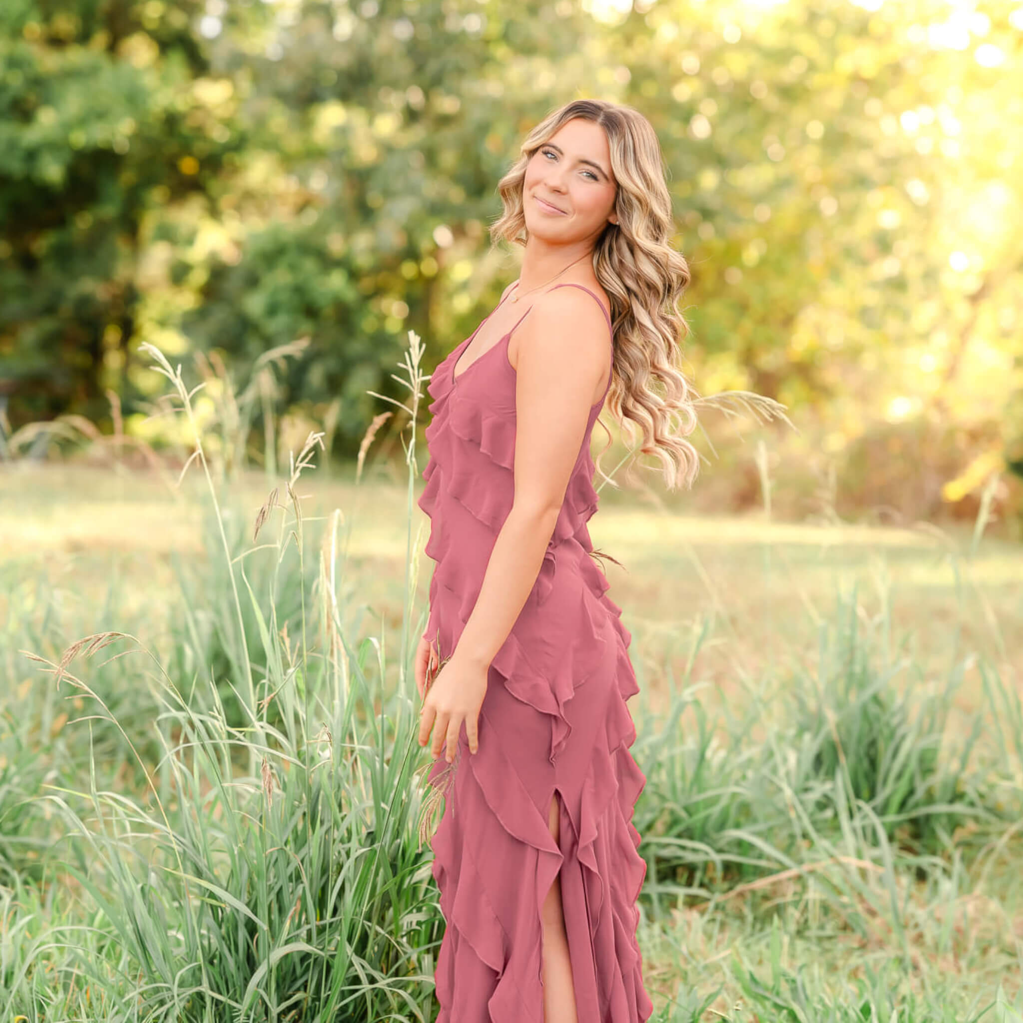 A high school senior, in a pink ruffled dress, tosses her hair over her should as she looks into the camera. The sun in shining through the trees behind her and the tall grass is green and wild.