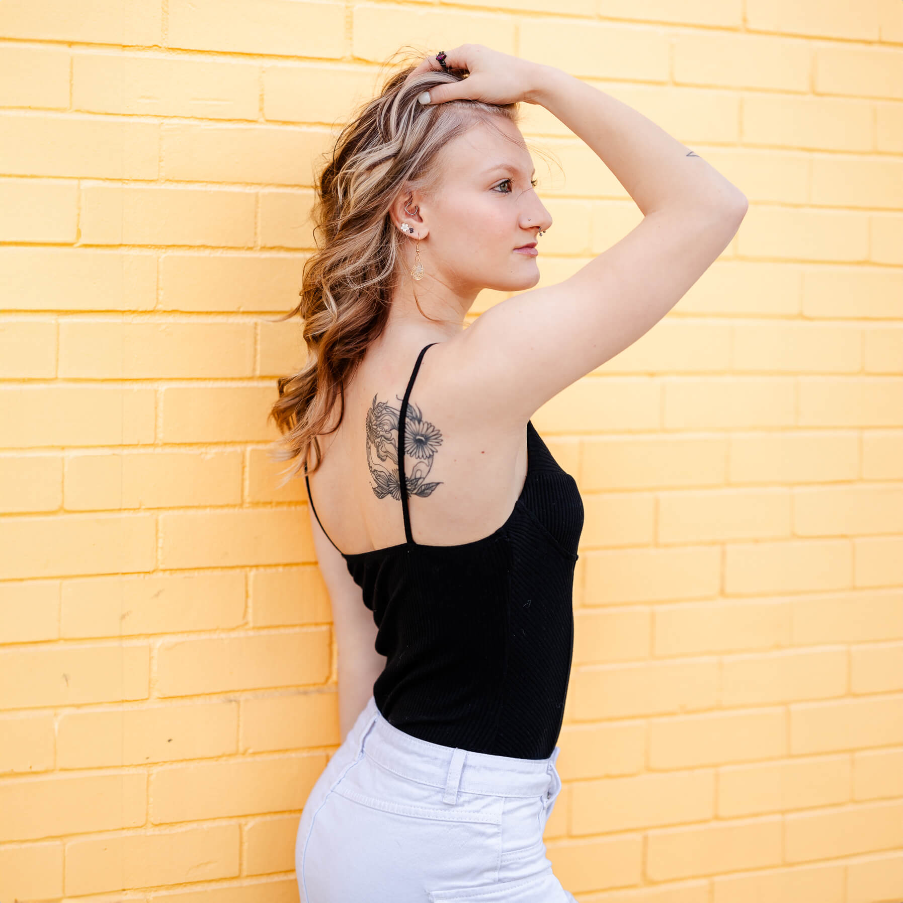 A high school senior shows off the tattoo on her shoulder. She is holding her hair up and looking off into the distance. There is a yellow, painted brick wall behind her.