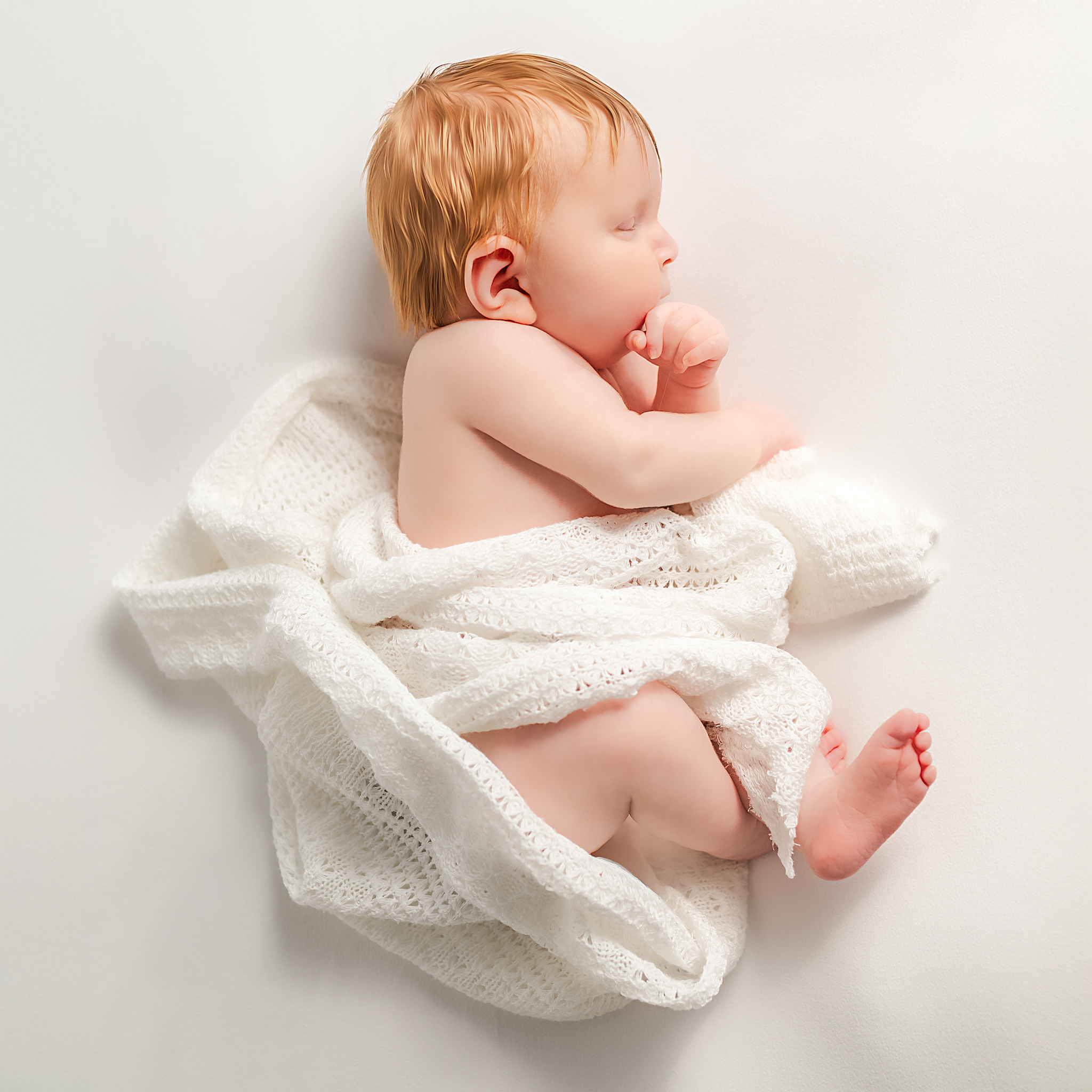 A newborn with red hair, sleeps on a white blanket, almost sucking her thumb. She is also wrapped in a white blanket.