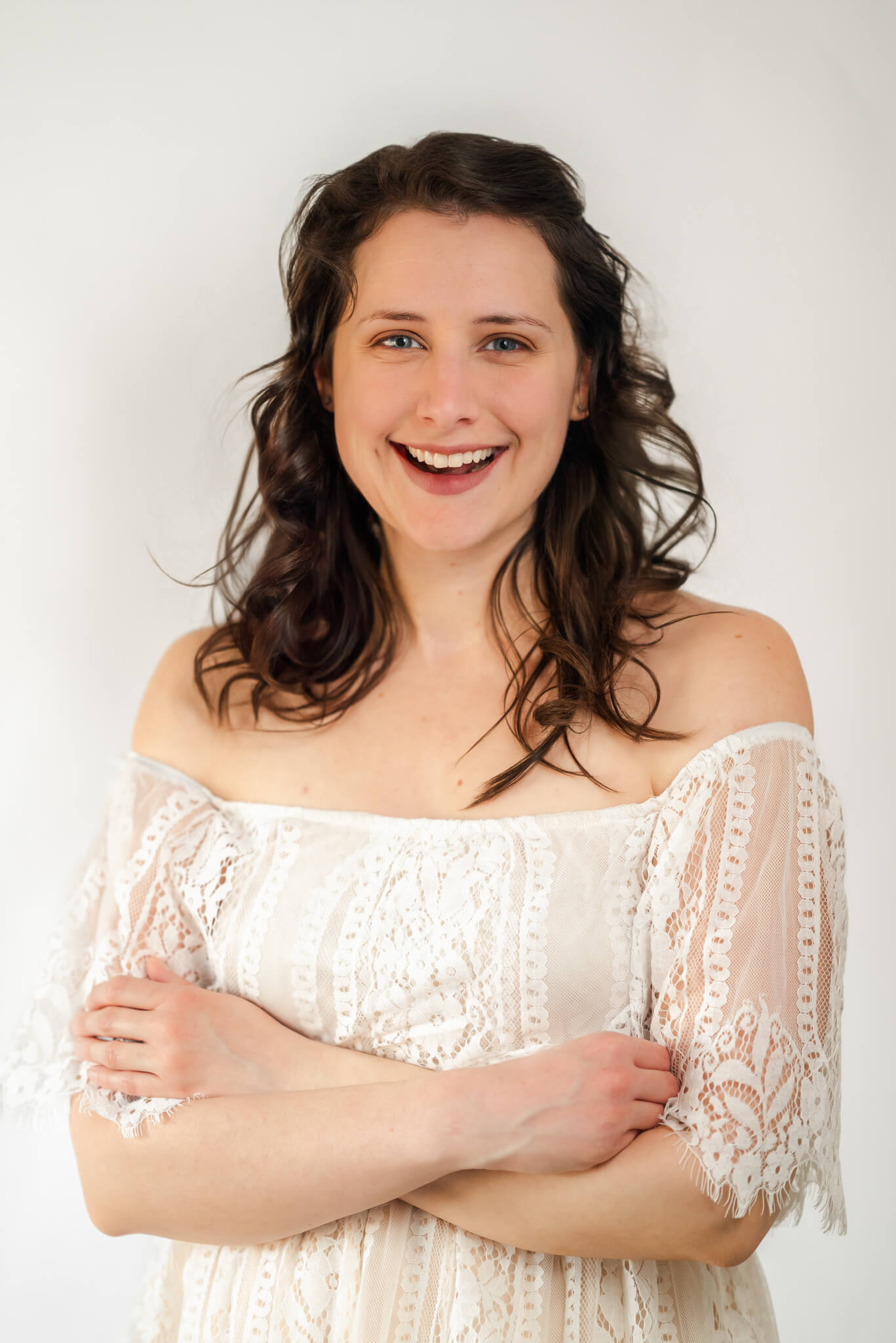 Image of the photographer at Justine Renee Photography. She is wearing a white lace dress and crossing her arms. There is a plain white background behind her.