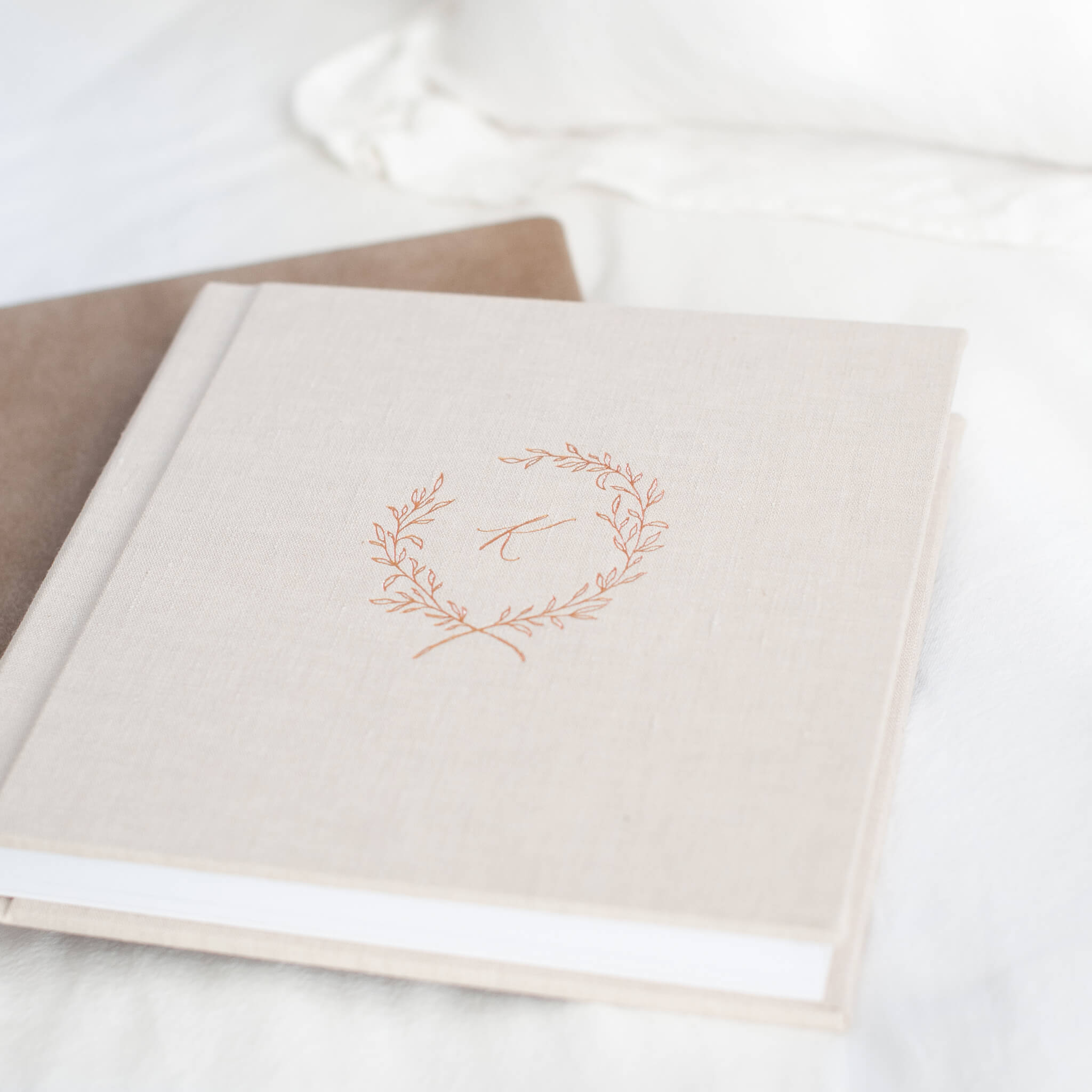 Two albums lay on a bed made up in white blankets and pillows. The top album is linen and debossed in rose gold.