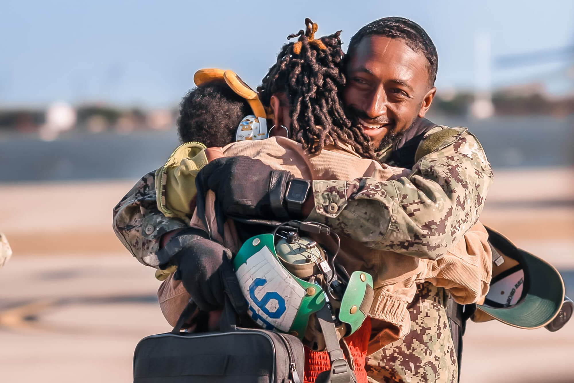 A sailor who has just returned from deployment hugs his young wife and baby son. He is smiling and holing back tears.