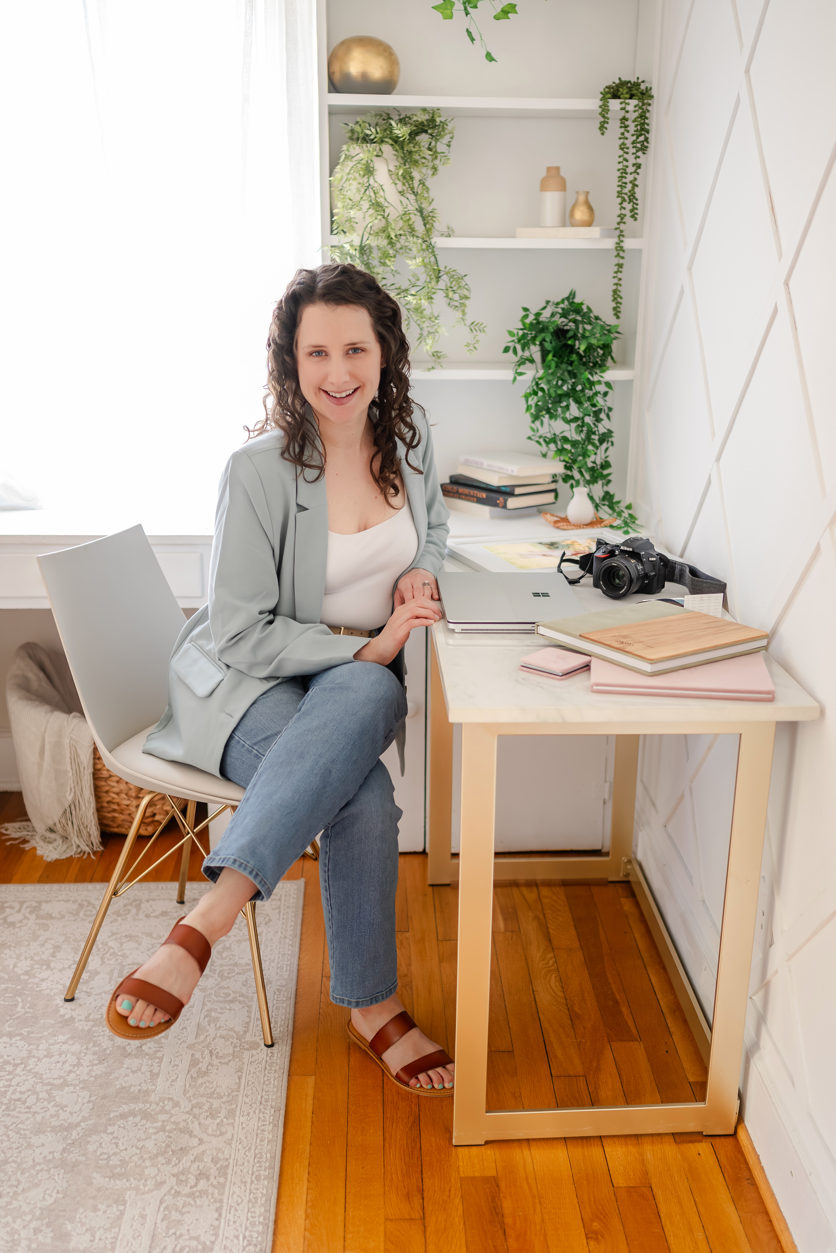 Image of Justine, the photographer at Justine Renee Photography. She is wearing a sage green blazer and blue jeans. She is sitting at a desk with her camera, laptop, and several product samples.