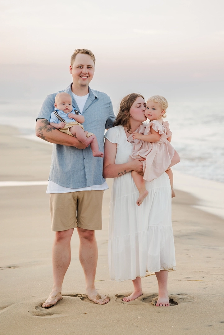 A family of four stands barefoot on the beach at sunset, with the father holding a baby boy and the mother kissing their toddler daughter on the cheek—capturing a sweet moment of Virginia Beach family photography against a pastel sky and gentle ocean waves.