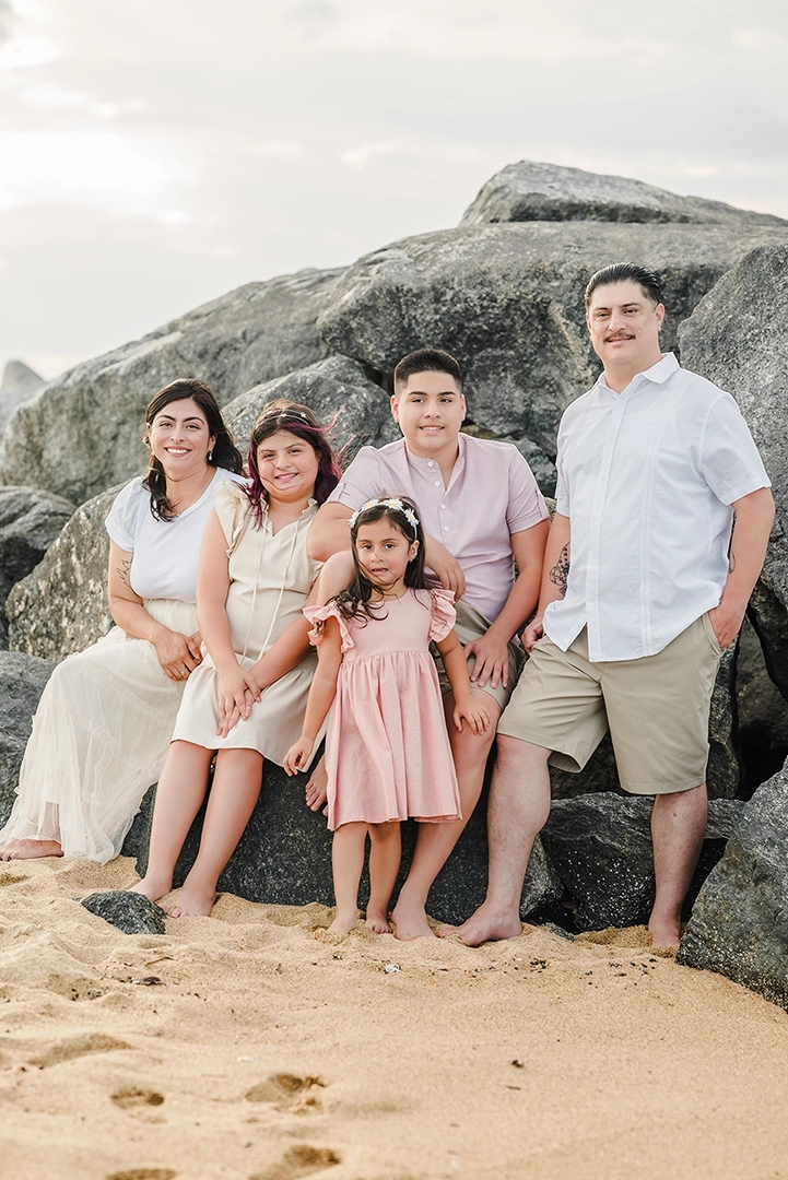 Family of five posing barefoot on a sandy beach in front of large rocks during a Virginia Beach photography session, dressed in coordinated pastel outfits with soft natural lighting.
