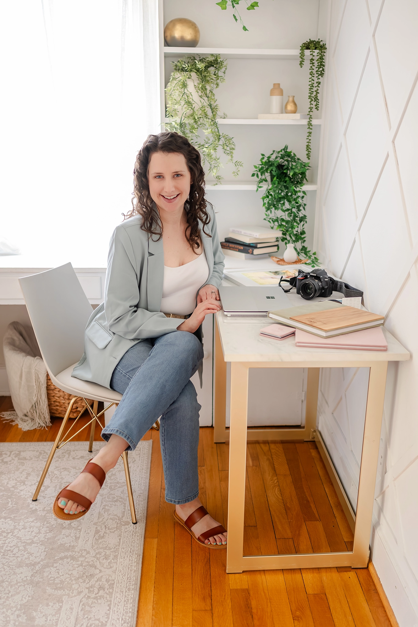 OBX photographer, Justine Renee Photography is pictured. She is wearing a sage green blazer and jeans. She is sitting at a white and gold desk with photo products and her camera spread out on it.