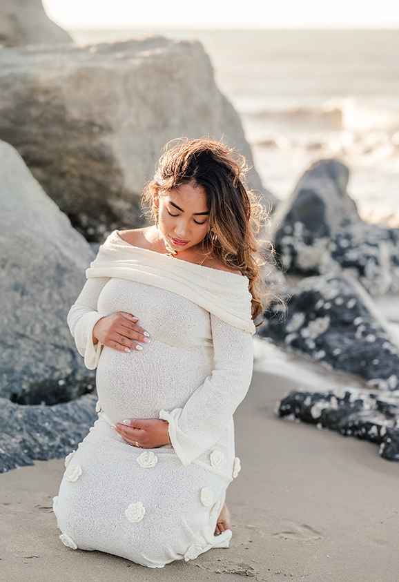 An expectant mother sits on the sand at the beach and holds her belly. She is wearing an off-white sweater dress and her hair is blowing in the wind. Image captured by OBX photographer, Justine Renee Photography.