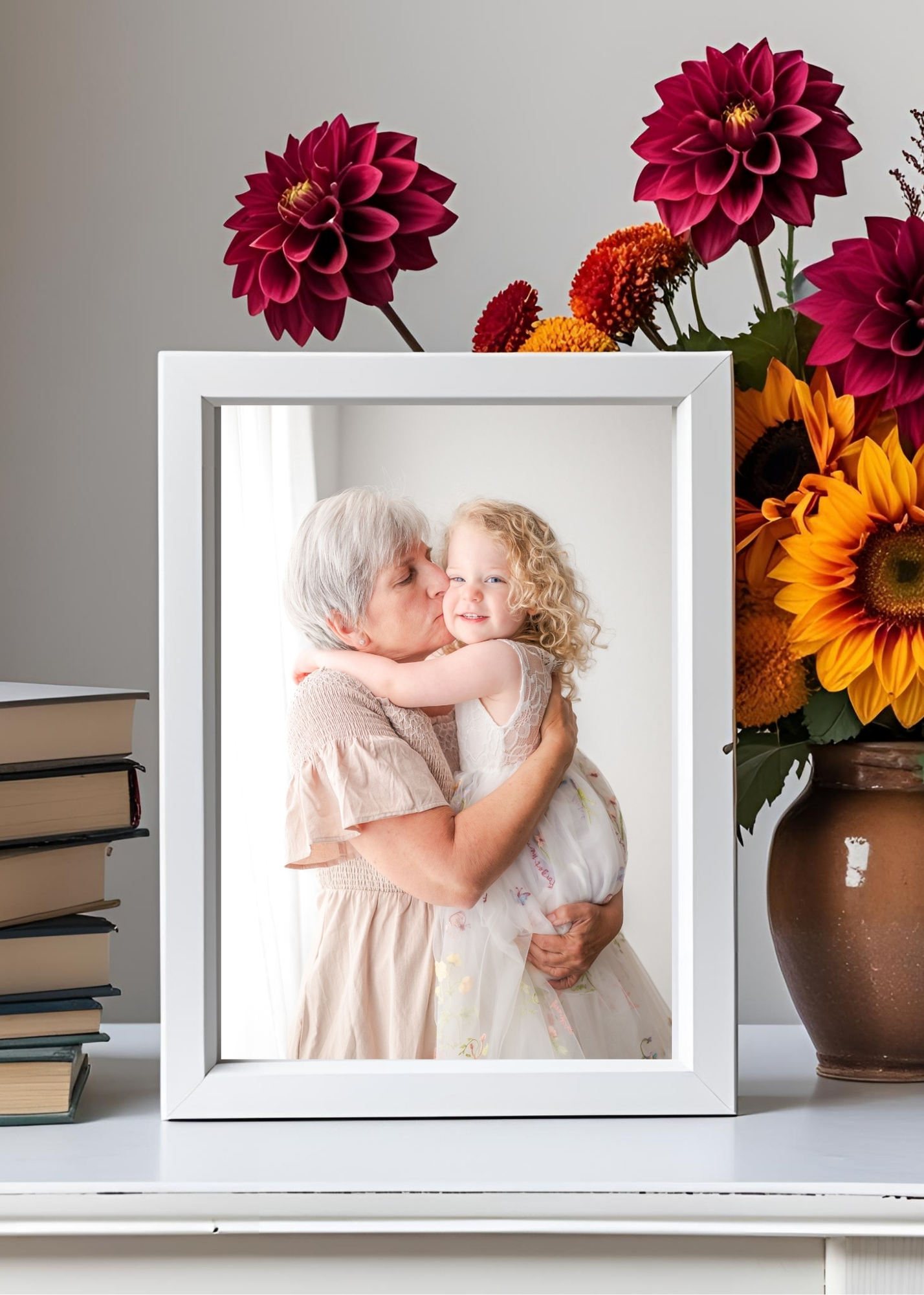 A desktop 5x7 frame surrounded by autumn blooms and a stack of books. The frame features and image of a toddler and her grandmother.