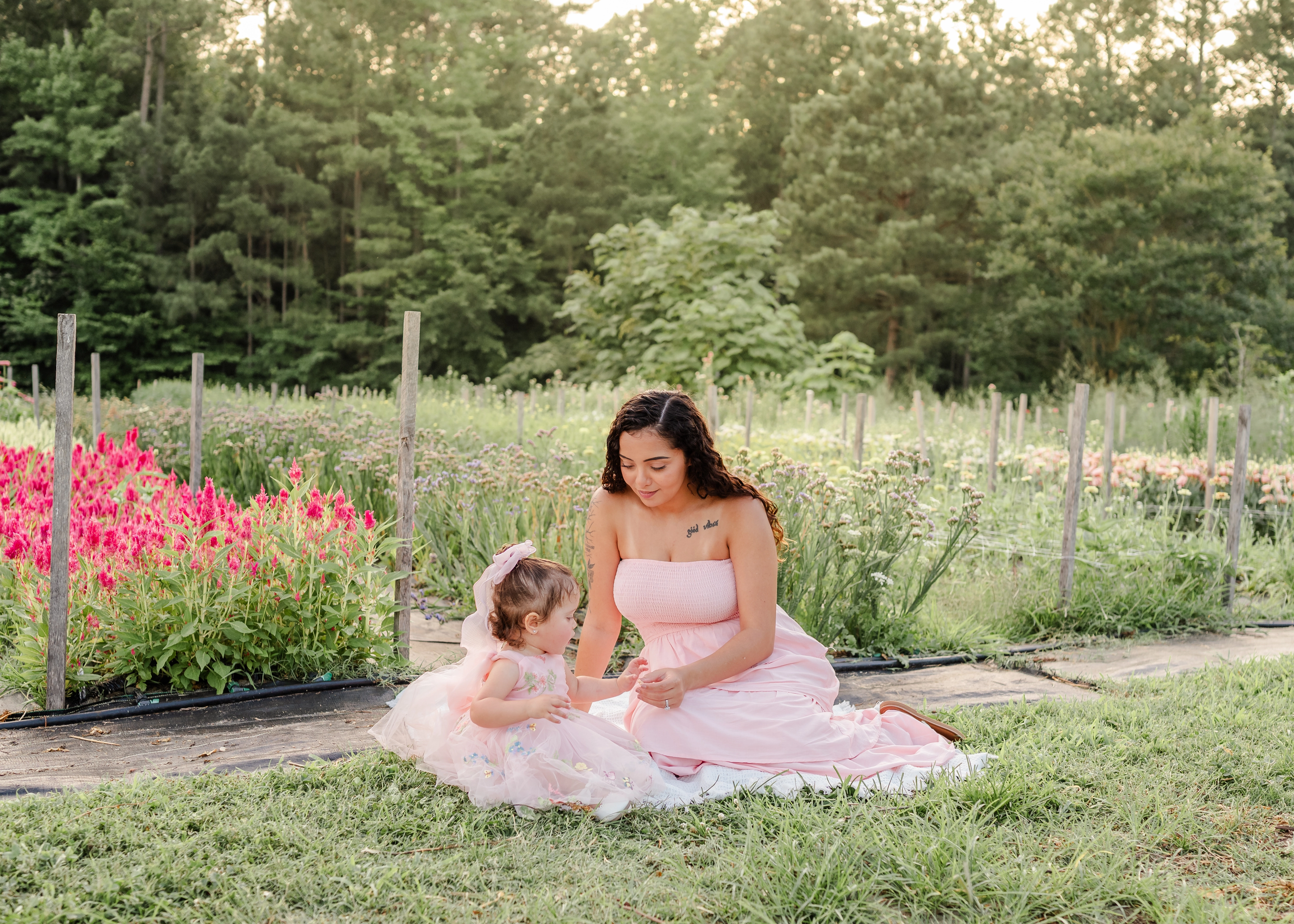 A mother in a light pink dress sits on a white blanket with her toddler, who is in a pink tulle dress. They are surrounded by flowers growing in rows and green trees behind the field.