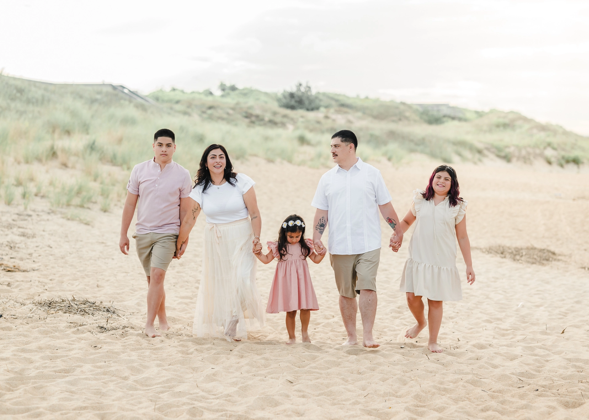 A family of 5 walks along the beach, with the grassy dunes behind them. They are wearing whites, pinks, and khakis.