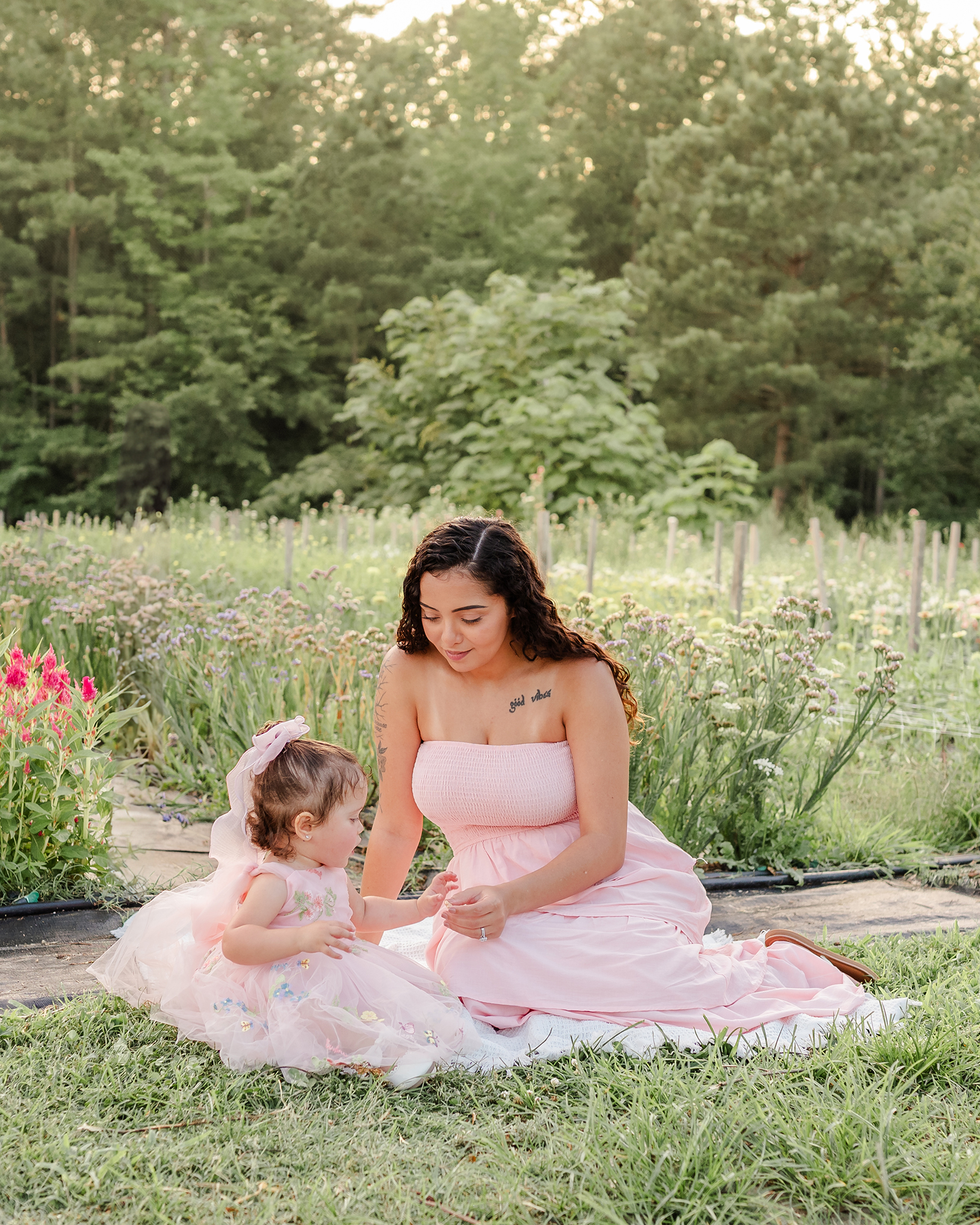 A mother and daughter, both in light pink dresses, sit on a blanket in the grass. Behind them are rows of flowers and the sun setting behind some trees.