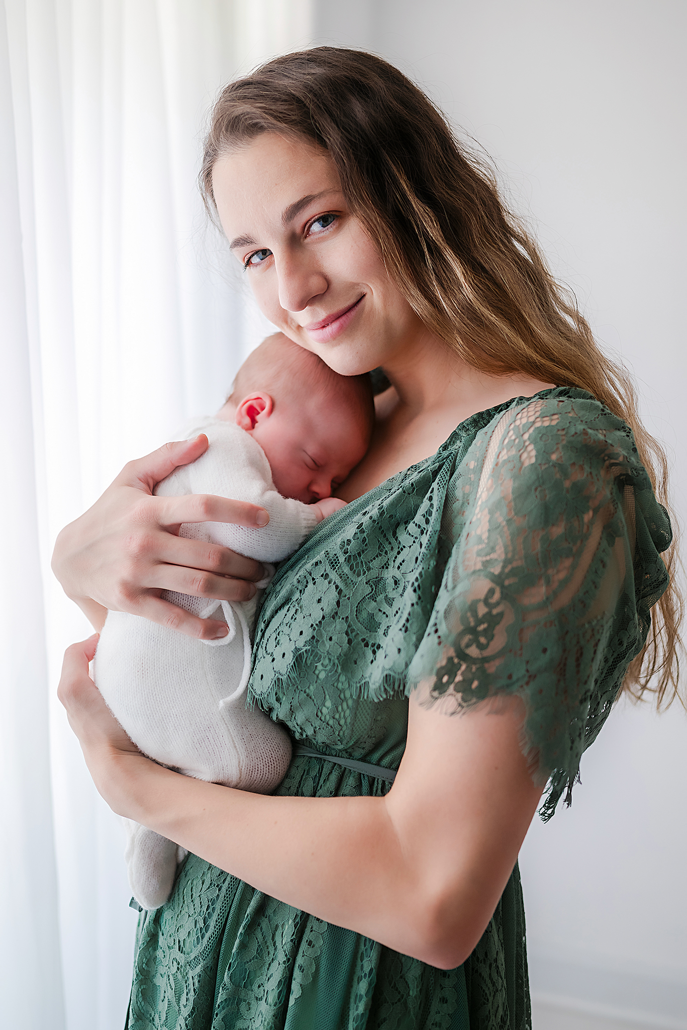 A young mother, holding her newborn baby, captured by Moyock photographer, Justine Renee Photography. The mother, who has long brown hair, is wearing a teal lace dress. Her son, who is snuggled on her chest, is in a white knitted romper.