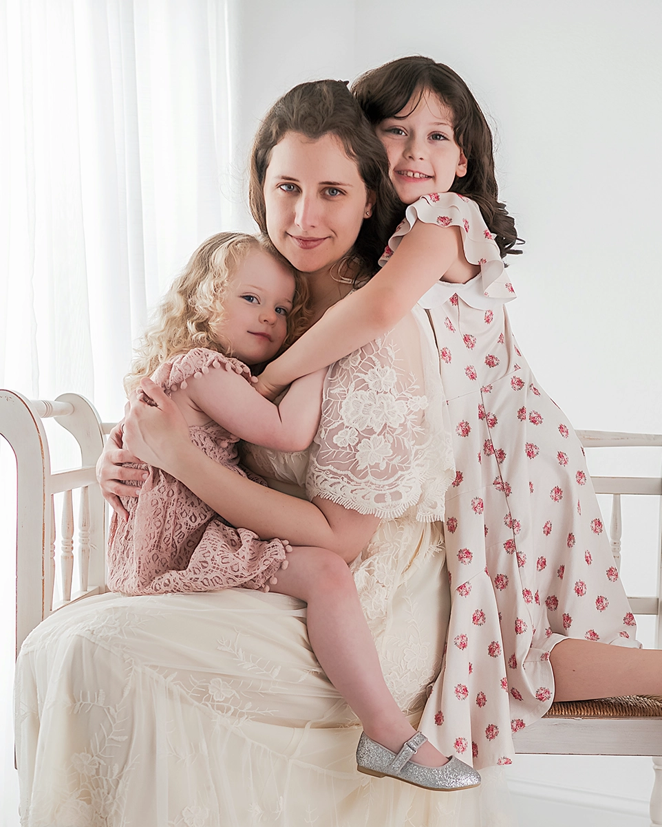 Justine, the photographer at Justine Renee Photography, is sitting on a bench wearing a white lace dress. Her toddler daughter, in pink, sit in her lap and rests her head on her chest Her older daughter in a white and pink floral dress, hugs her from behind.