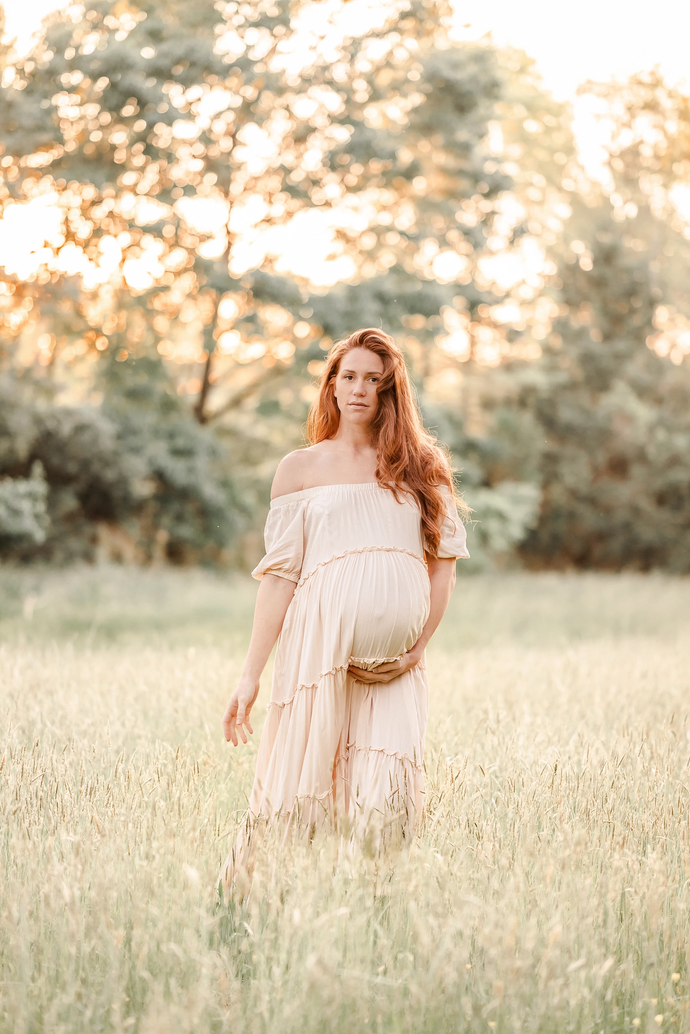 An expectant mother stands in a field of tall grass during her maternity session with Chesapeake photographer, Justine Renee Photography. She is wearing a flowy beige dress and the sun is shining through the trees behind her.