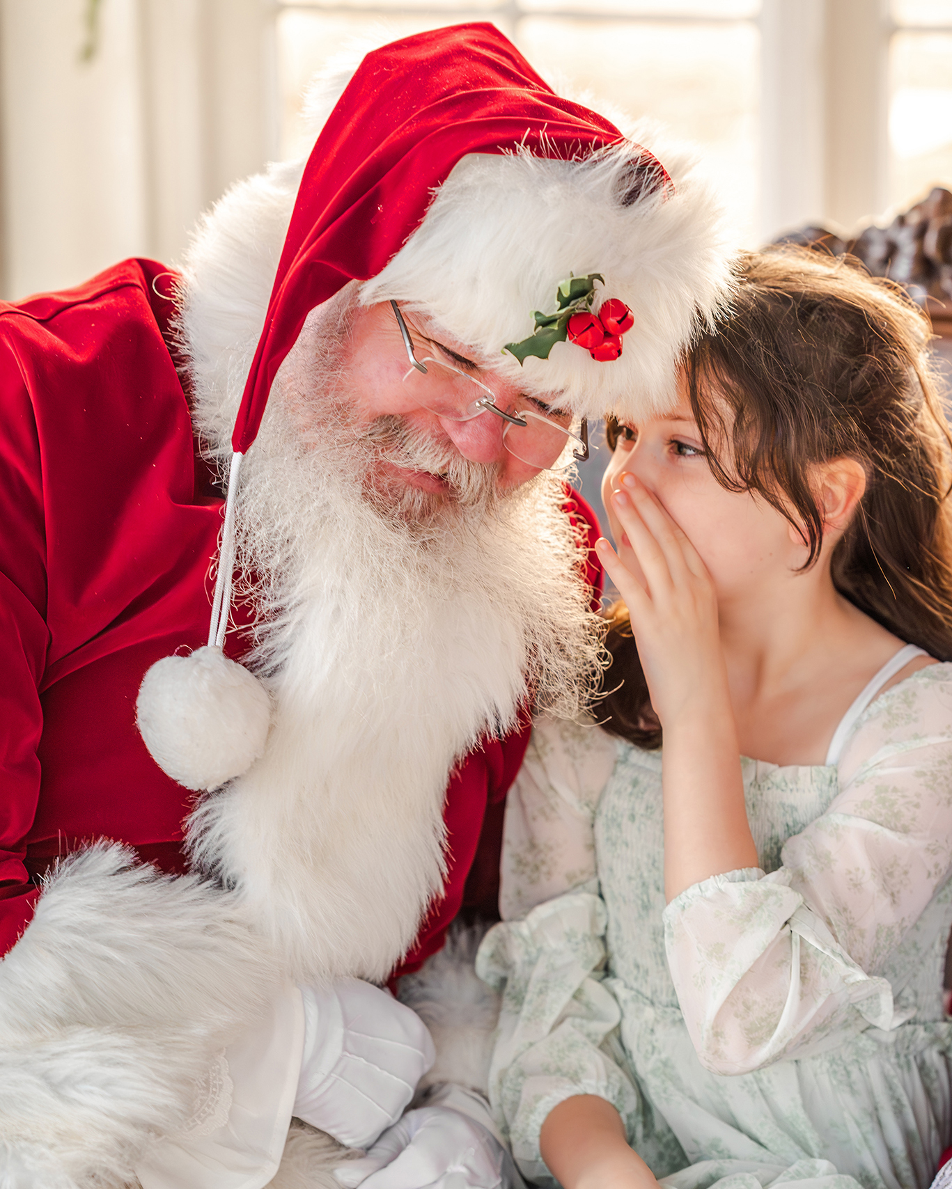 A young girl, in a white and green dress, whispers in Santa's ear.