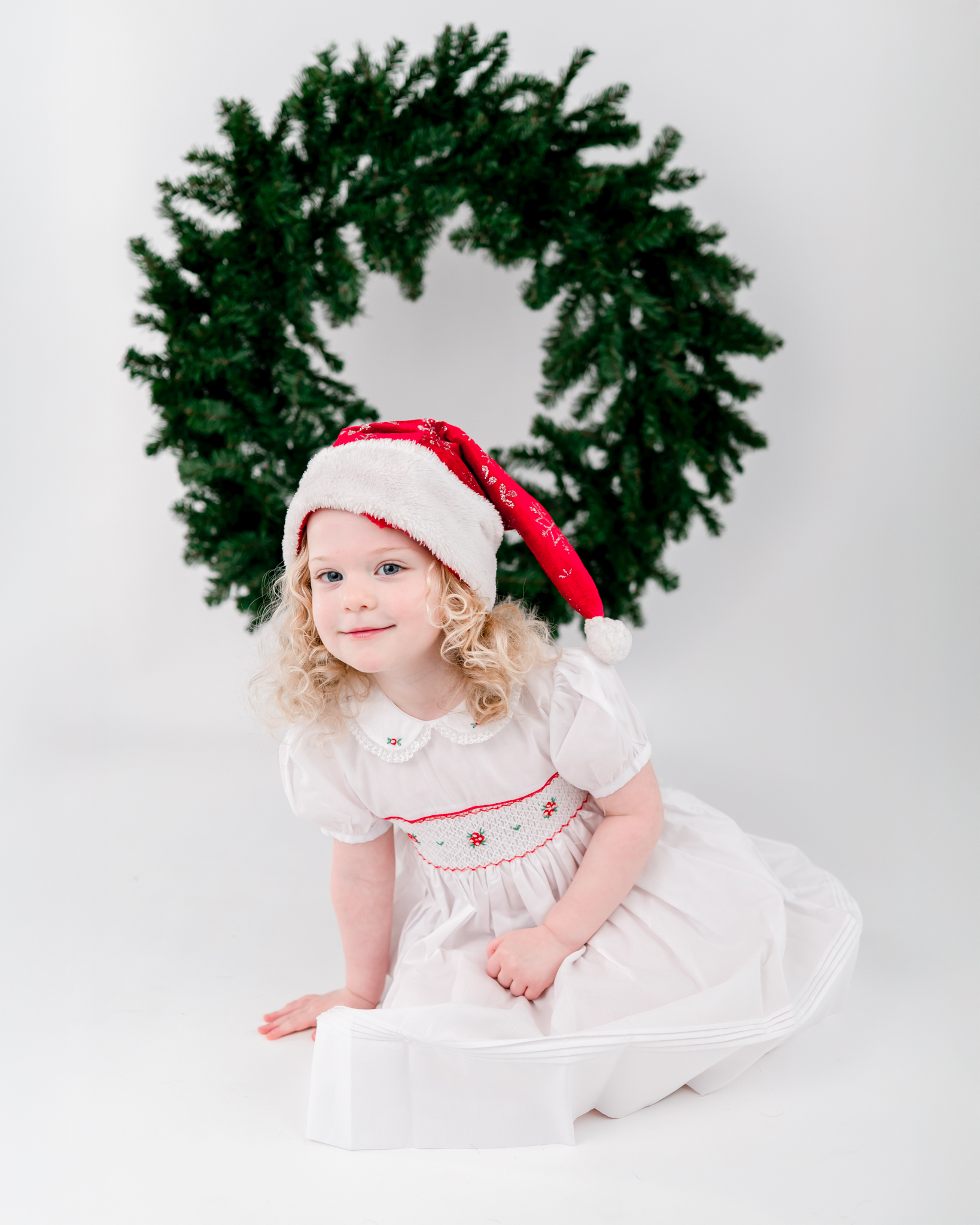 A toddler in a white dress and Santa cap, sits in front of a wreath on a white backdrop.