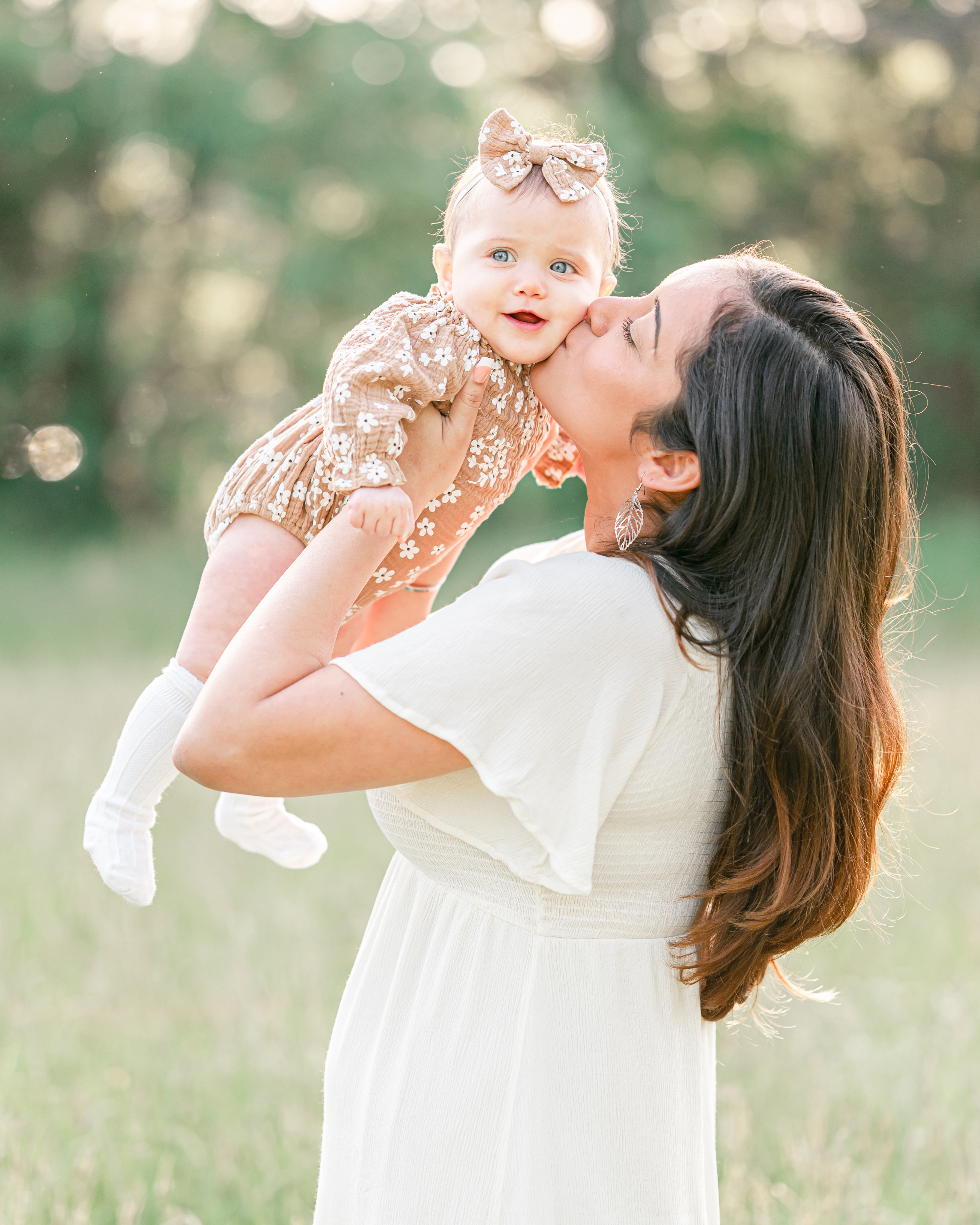 A mother in a white dress holds her toddler and kisses her cheek. The toddler is wearing a brown floral romper, which compliments the grassy field they are standing in.