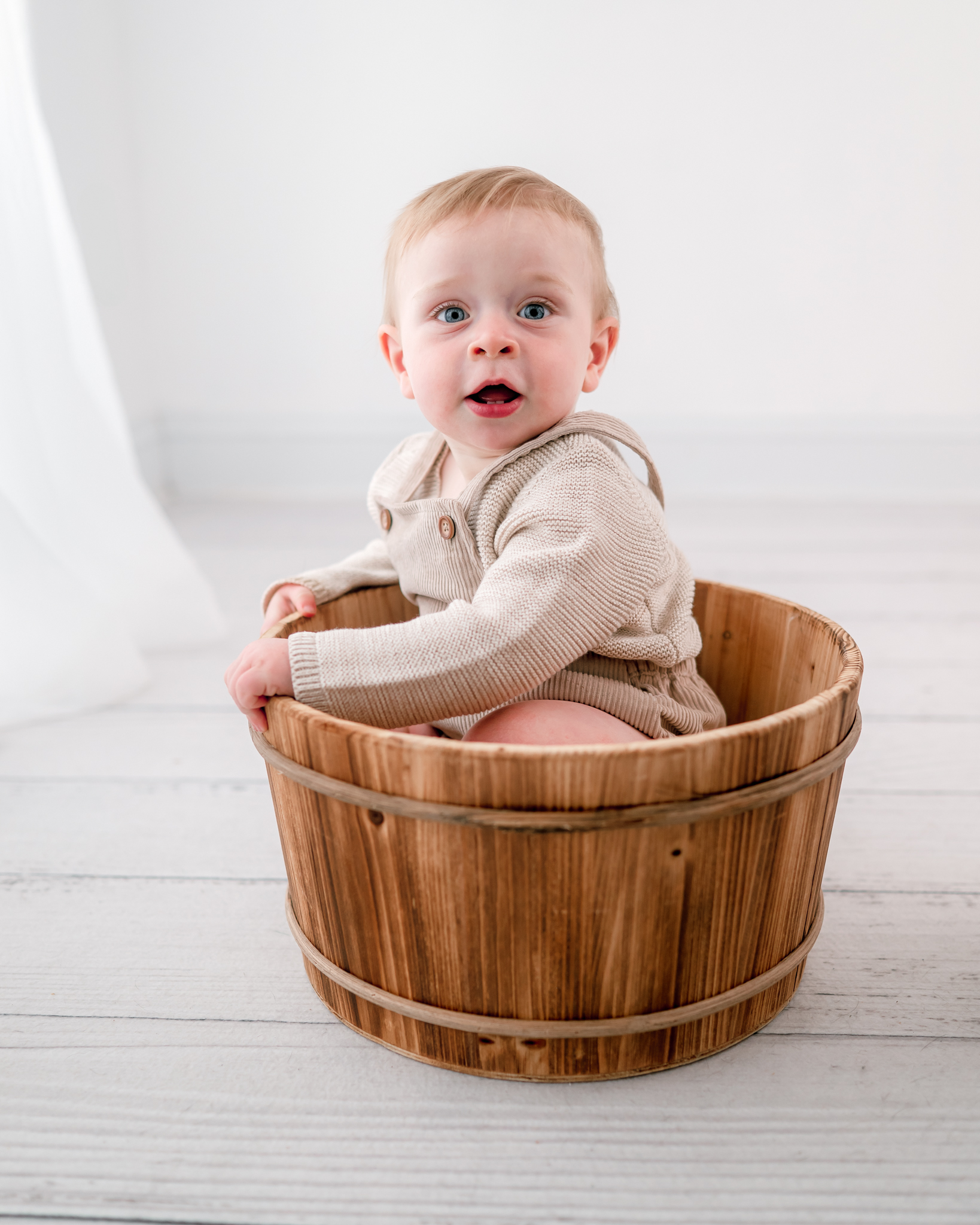 A 9 month old baby boy, wearing a beige overalls outfit, sits in a wooden bucket in a white room. He is looking right at the camera.