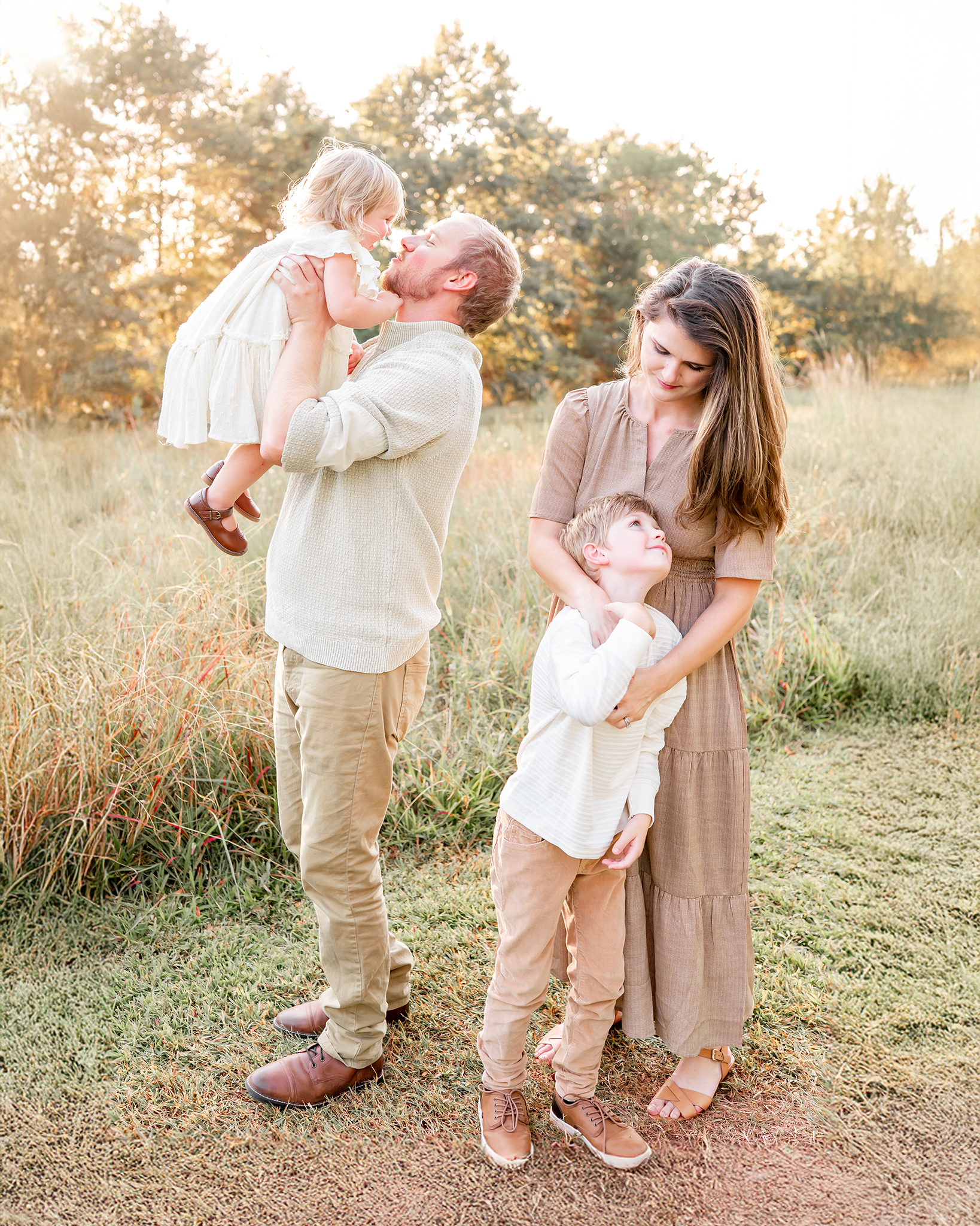 A mother in a white dress holds her toddler and kisses her cheek. The toddler is wearing a brown floral romper, which compliments the grassy field they are standing in.